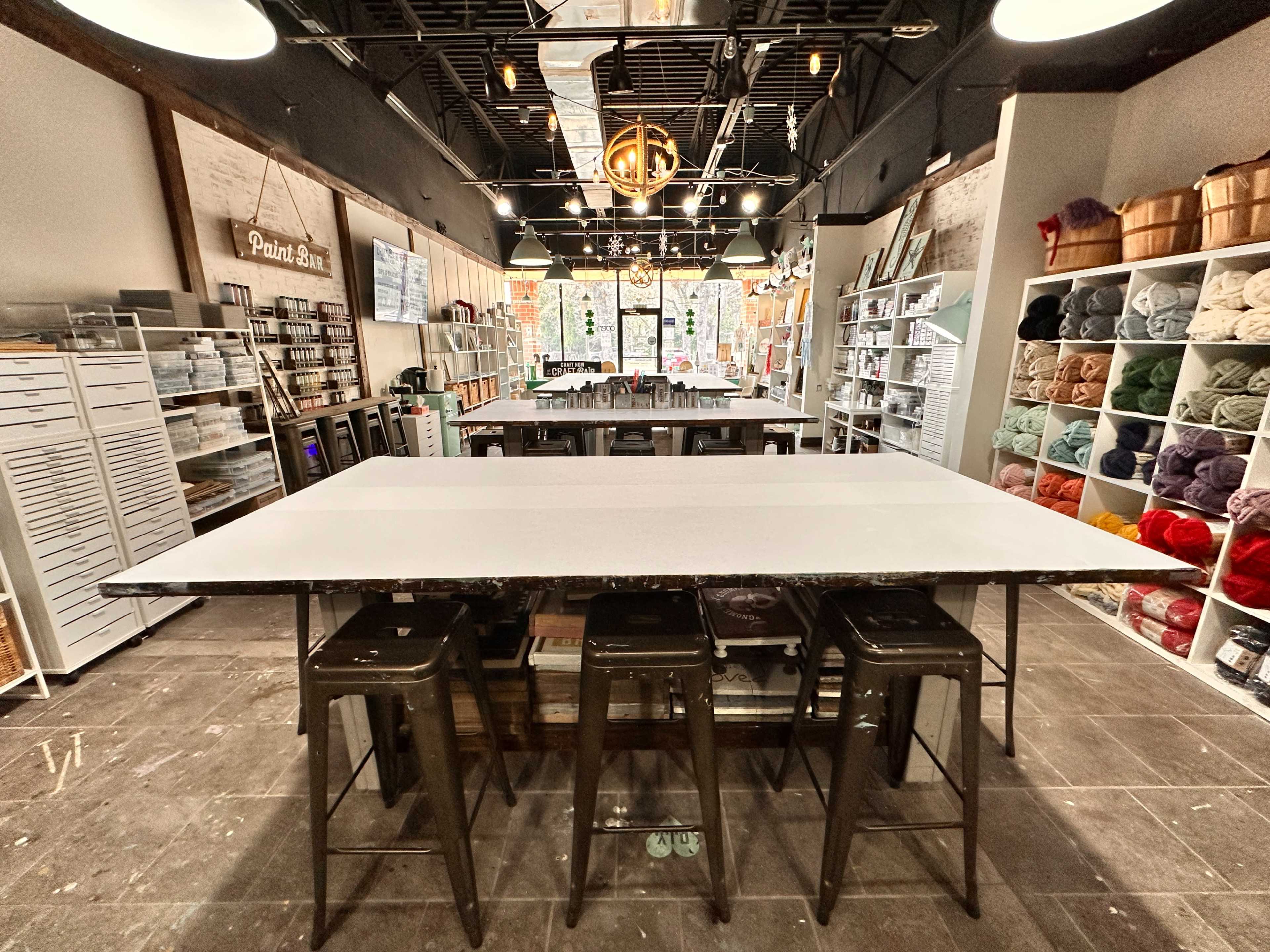 The image shows a spacious craft store interior featuring a central work table surrounded by shelves stocked with various art supplies and yarns.