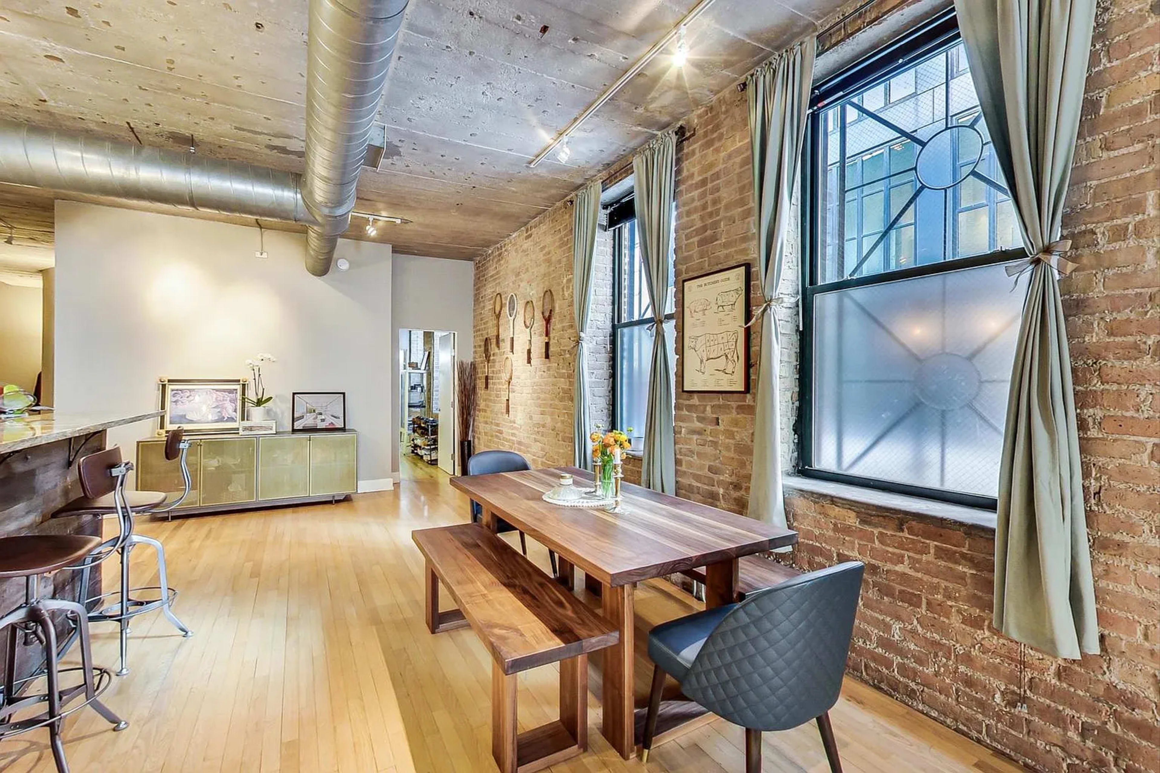 The image shows a modern dining area with a wooden table, chairs, and large windows featuring exposed brick walls.