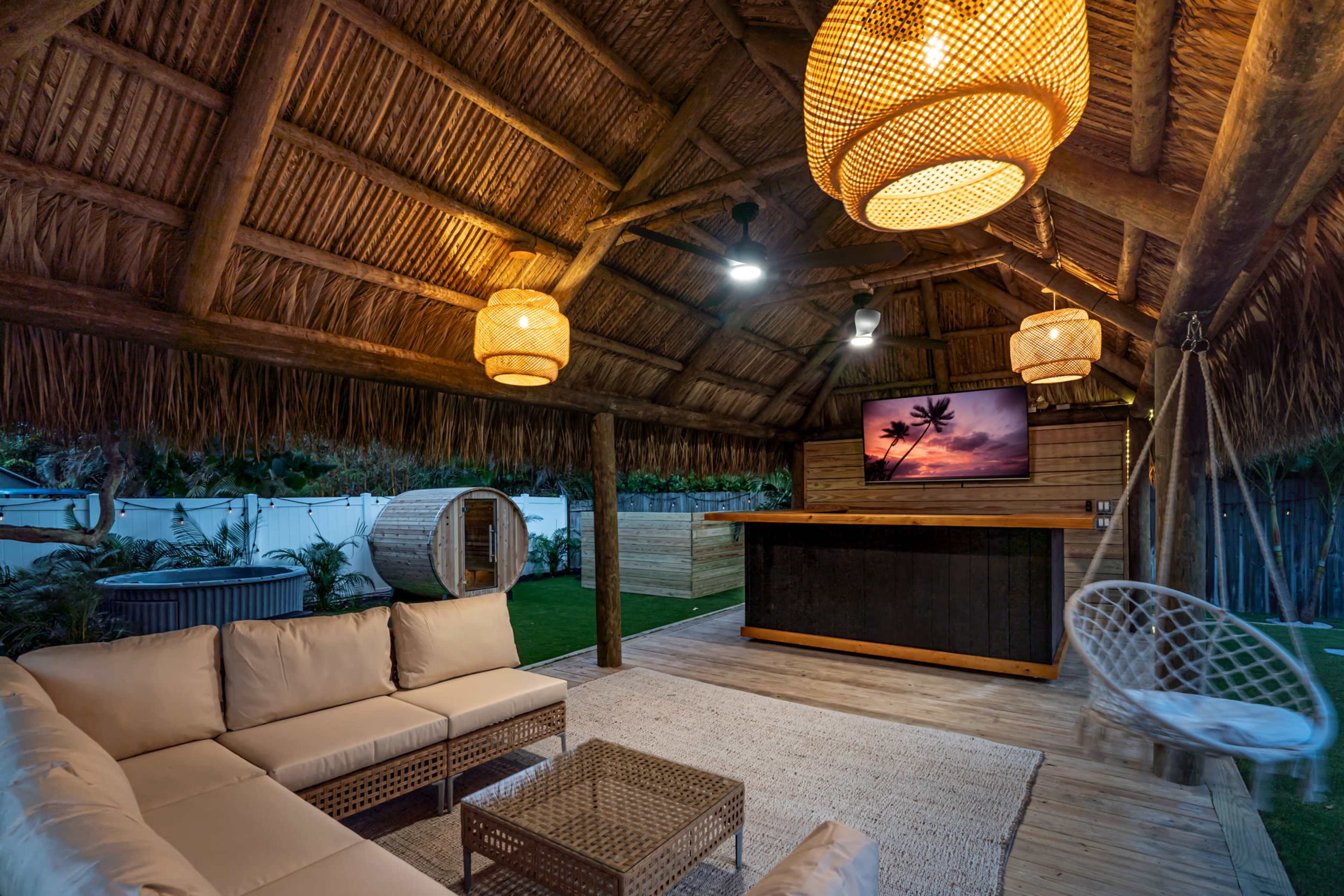 A rustic outdoor lounge area features a thatched roof, comfortable seating, a wooden bar, a television displaying a sunset scene, and decorative lighting.