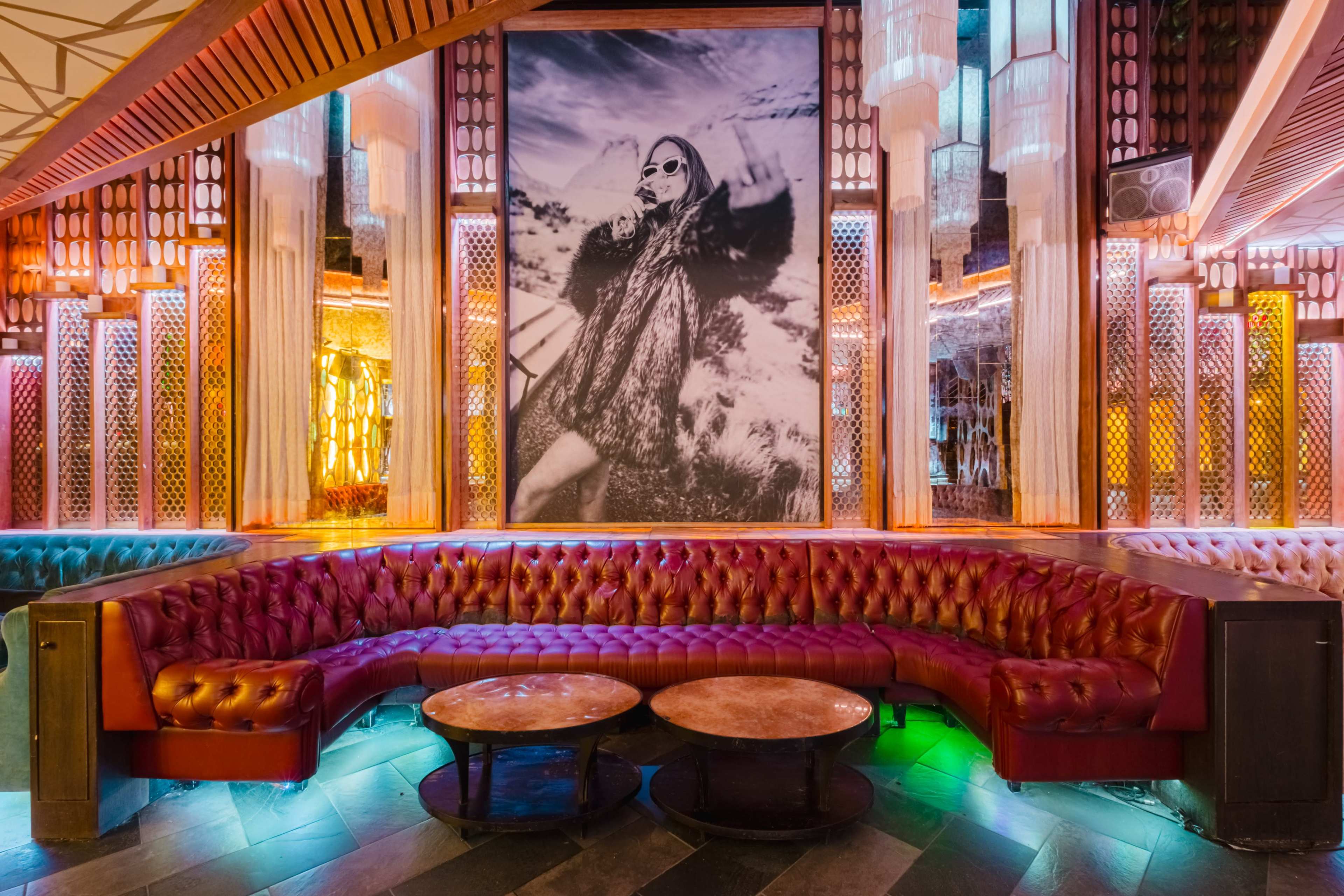 The image shows a stylish lounge area with a large black-and-white photograph on the wall, featuring a woman in a vintage outfit, surrounded by vibrant lighting and a red leather sectional couch.