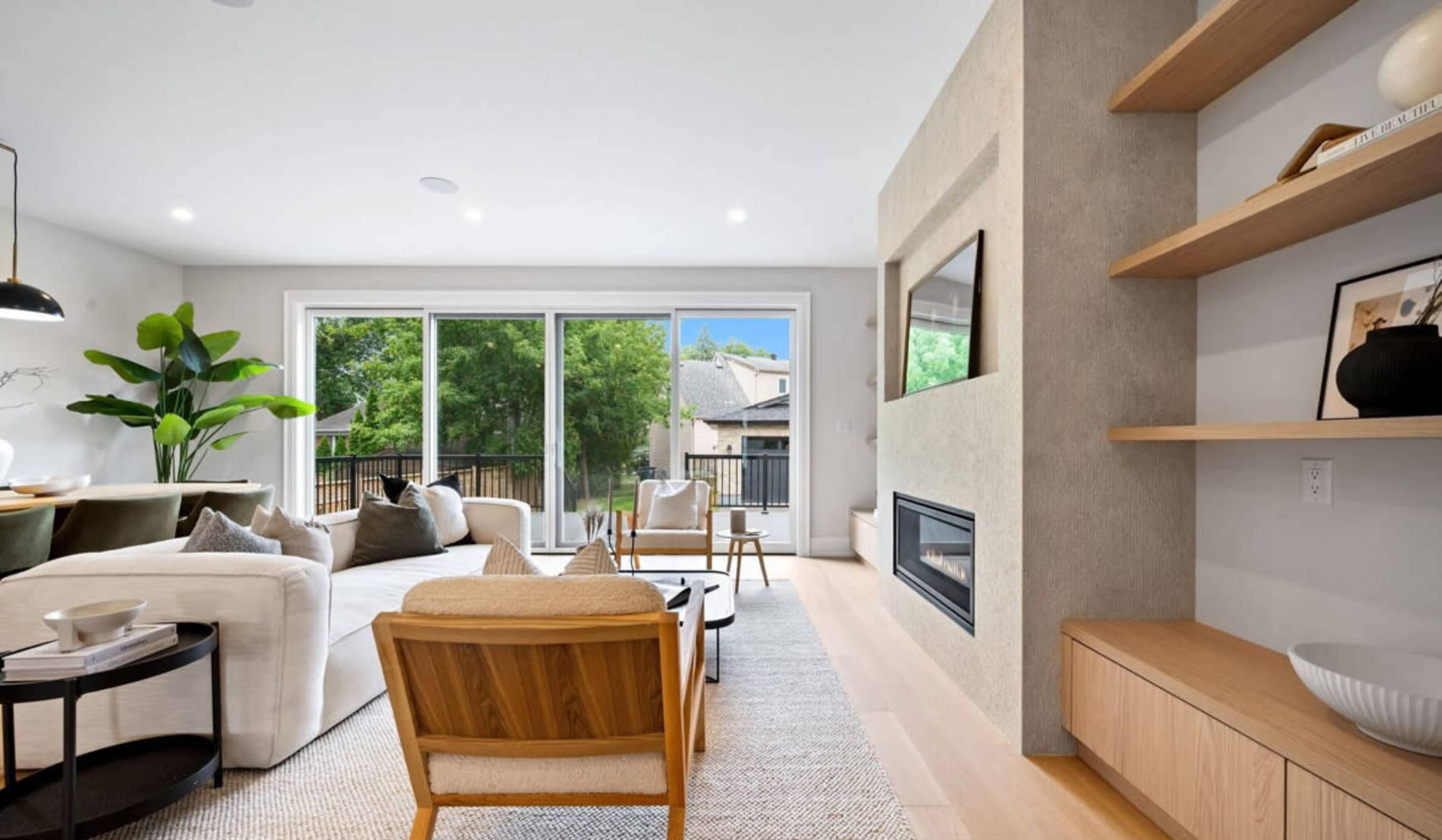 The image shows a modern living room with large windows overlooking greenery, a fireplace, and minimalist furniture.