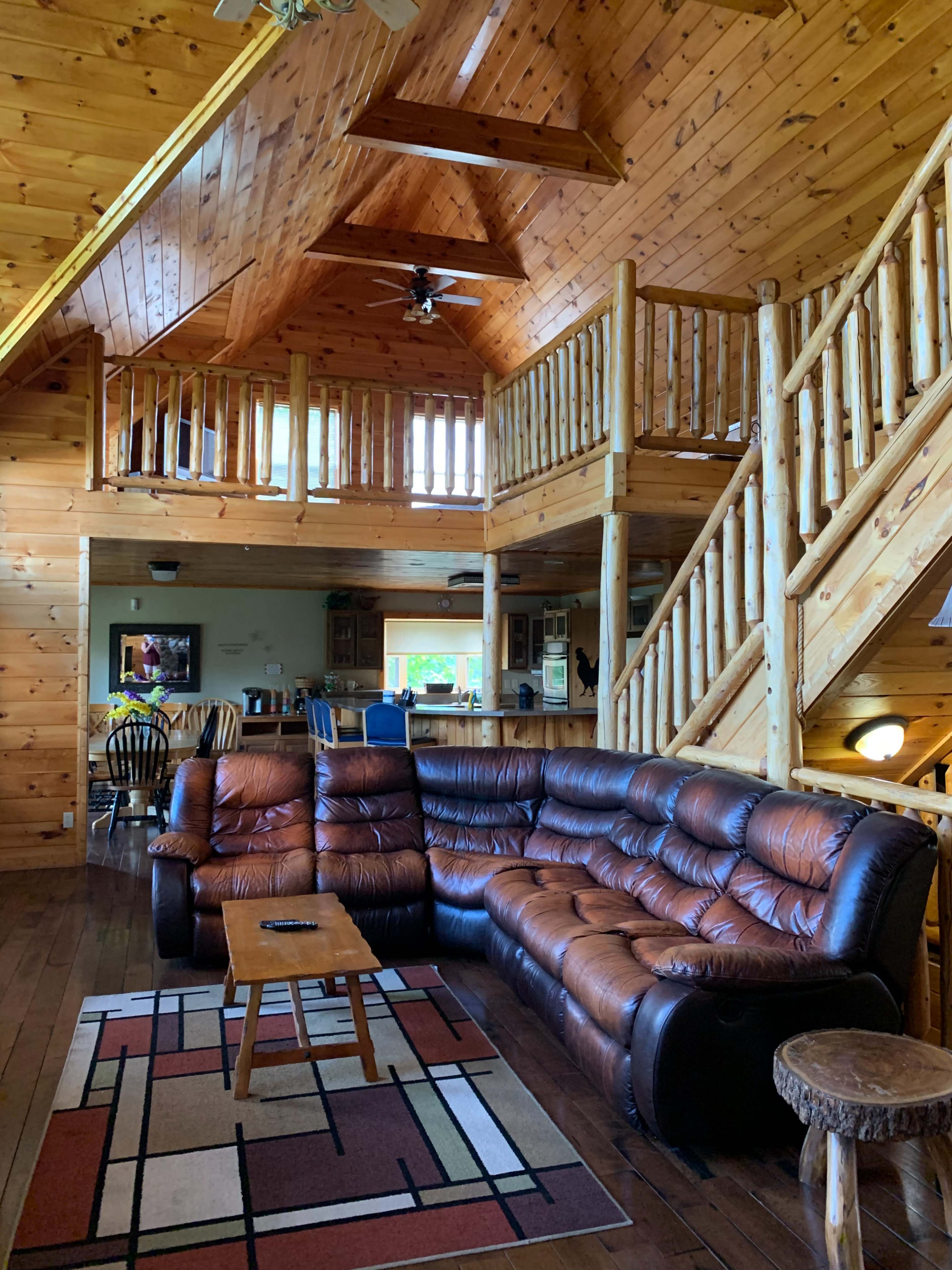 A spacious wooden interior of a cabin featuring a brown leather sectional sofa, a wooden coffee table, and an open staircase leading to a loft area.