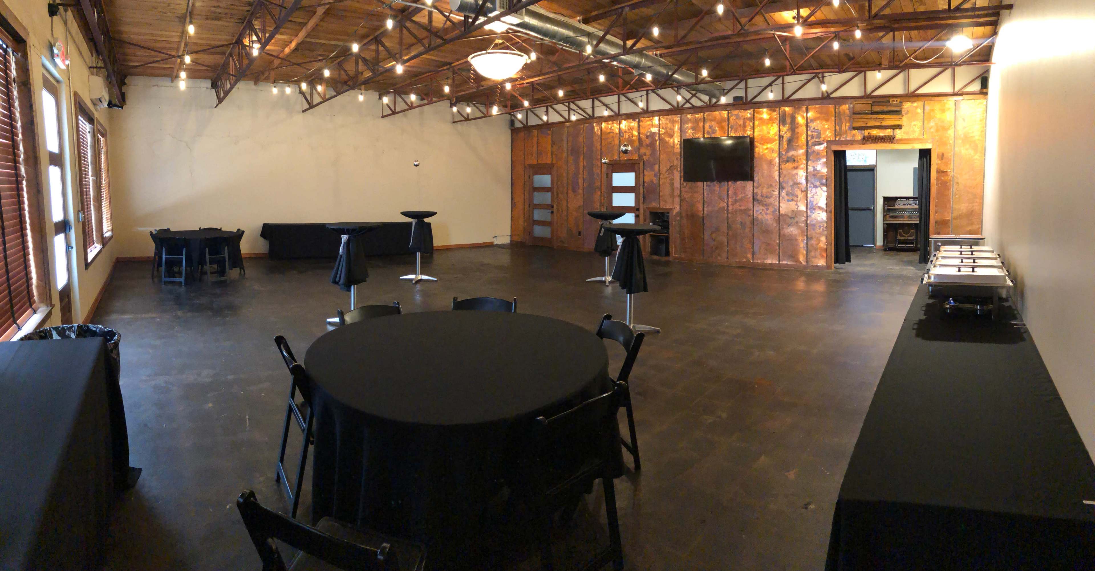 A spacious event room with dark flooring, a copper accent wall, and several tables and stands set up for an event.