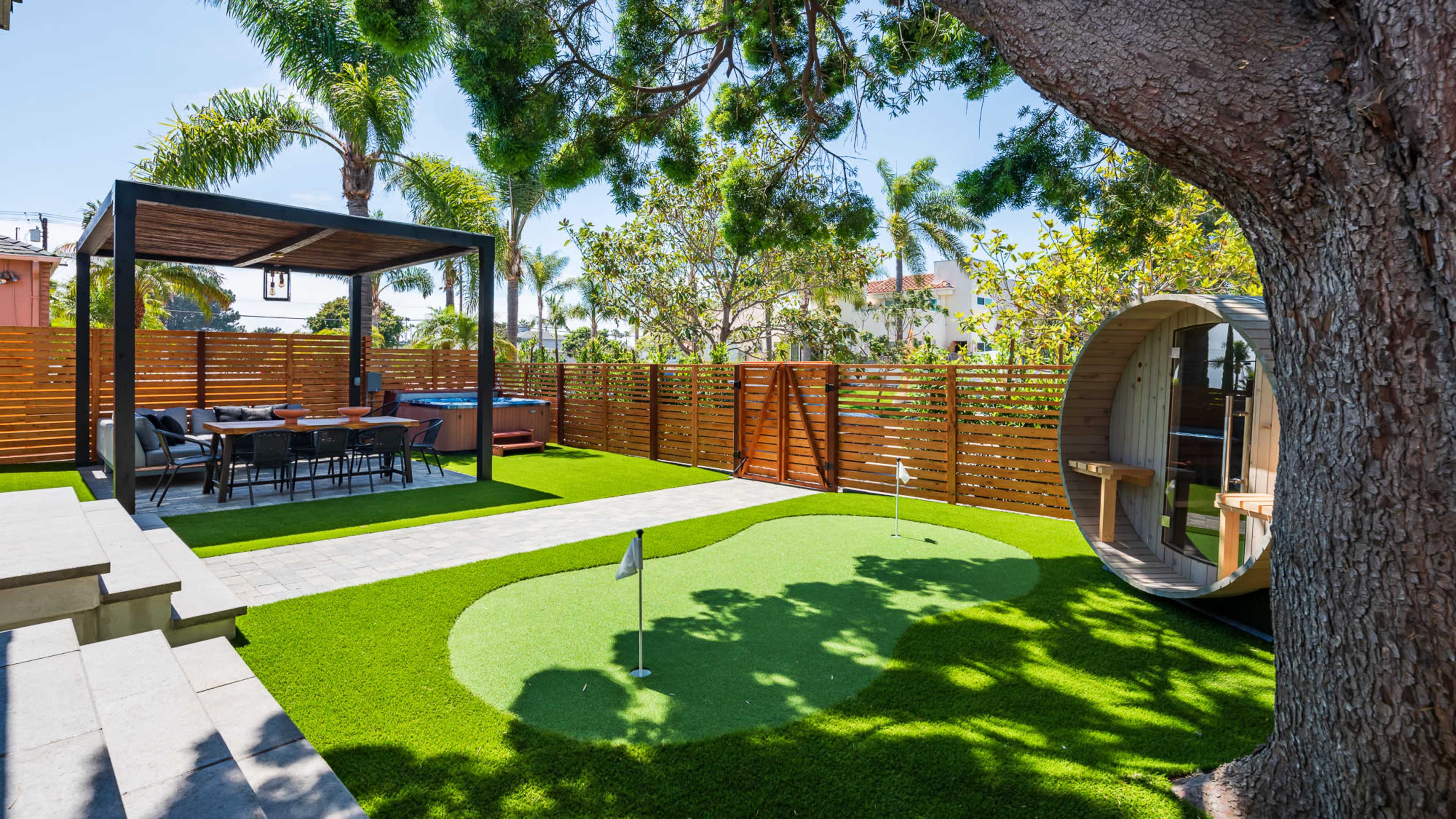 The image shows a landscaped backyard featuring a putting green, a dining area under a pergola, a hot tub, and a round wooden structure surrounded by palm trees.