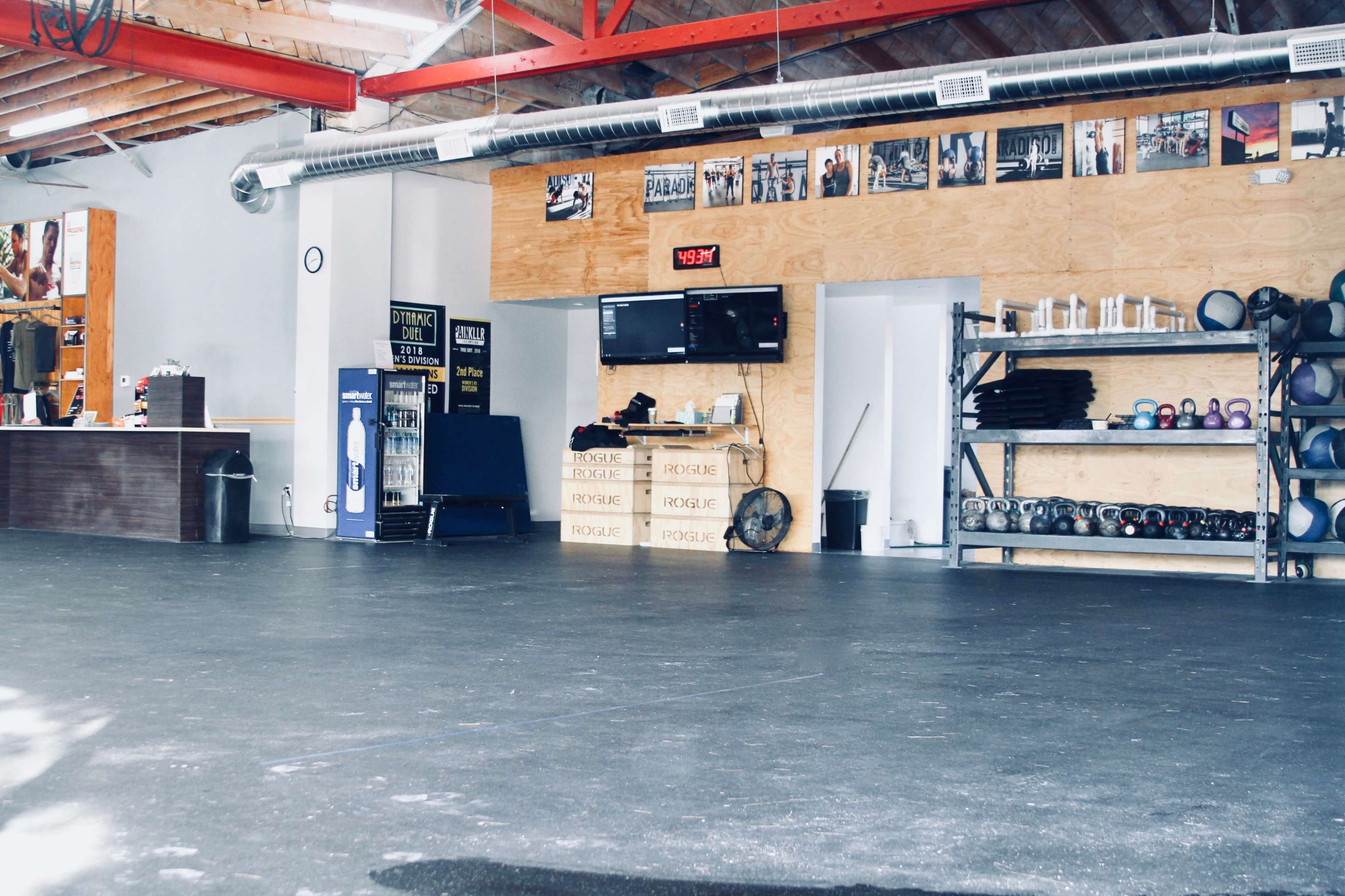 A spacious gym interior with wooden walls, workout equipment on shelves, a digital clock, and fitness posters on the walls.