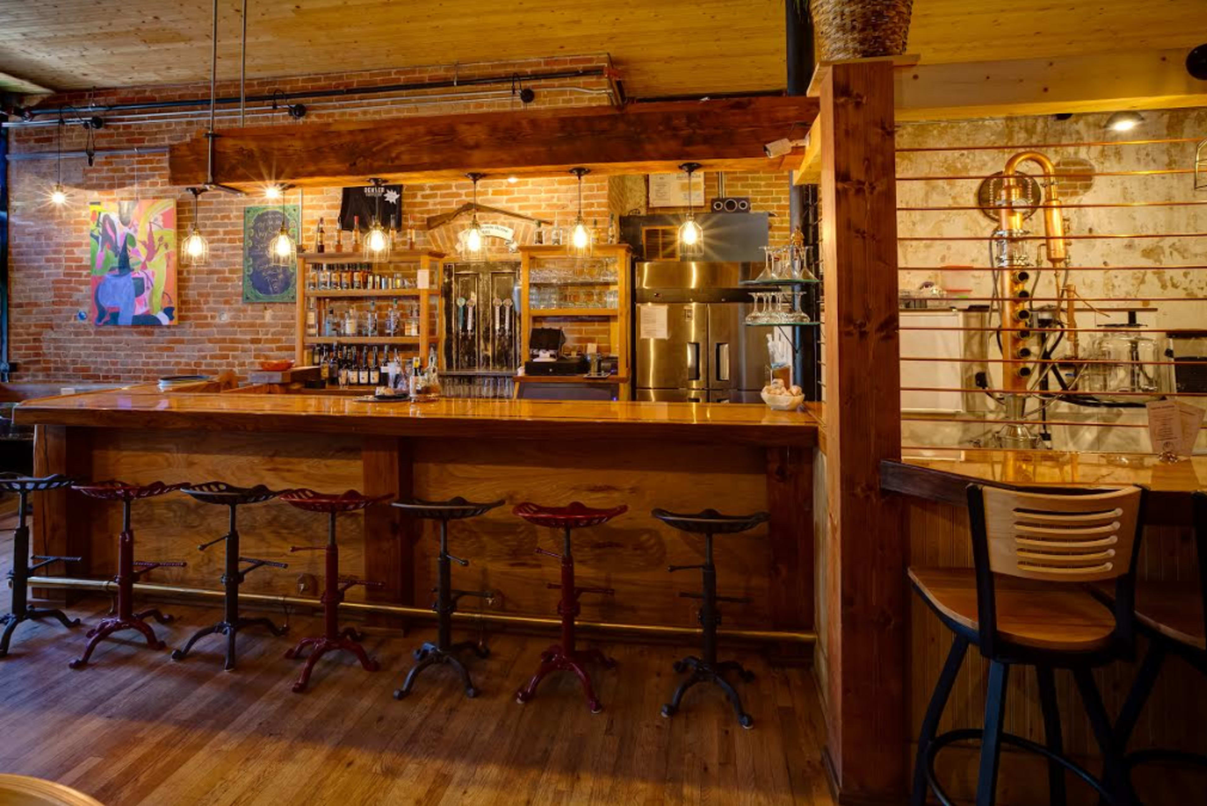 A rustic bar area with wooden counters, metal stools, and exposed brick walls, featuring shelves stocked with bottles of various beverages.