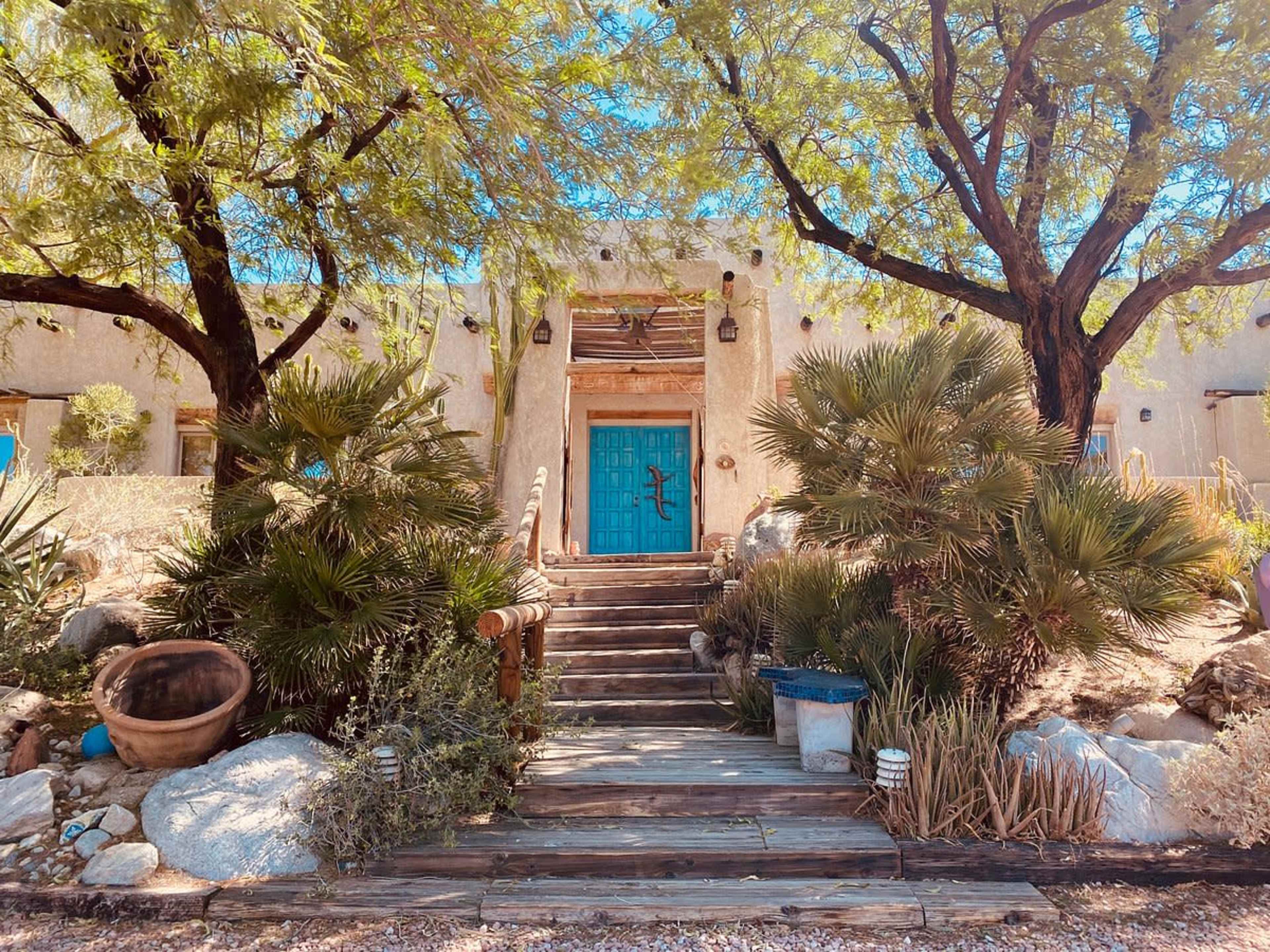 A staircase leads to a house with a blue door, surrounded by desert vegetation and trees.