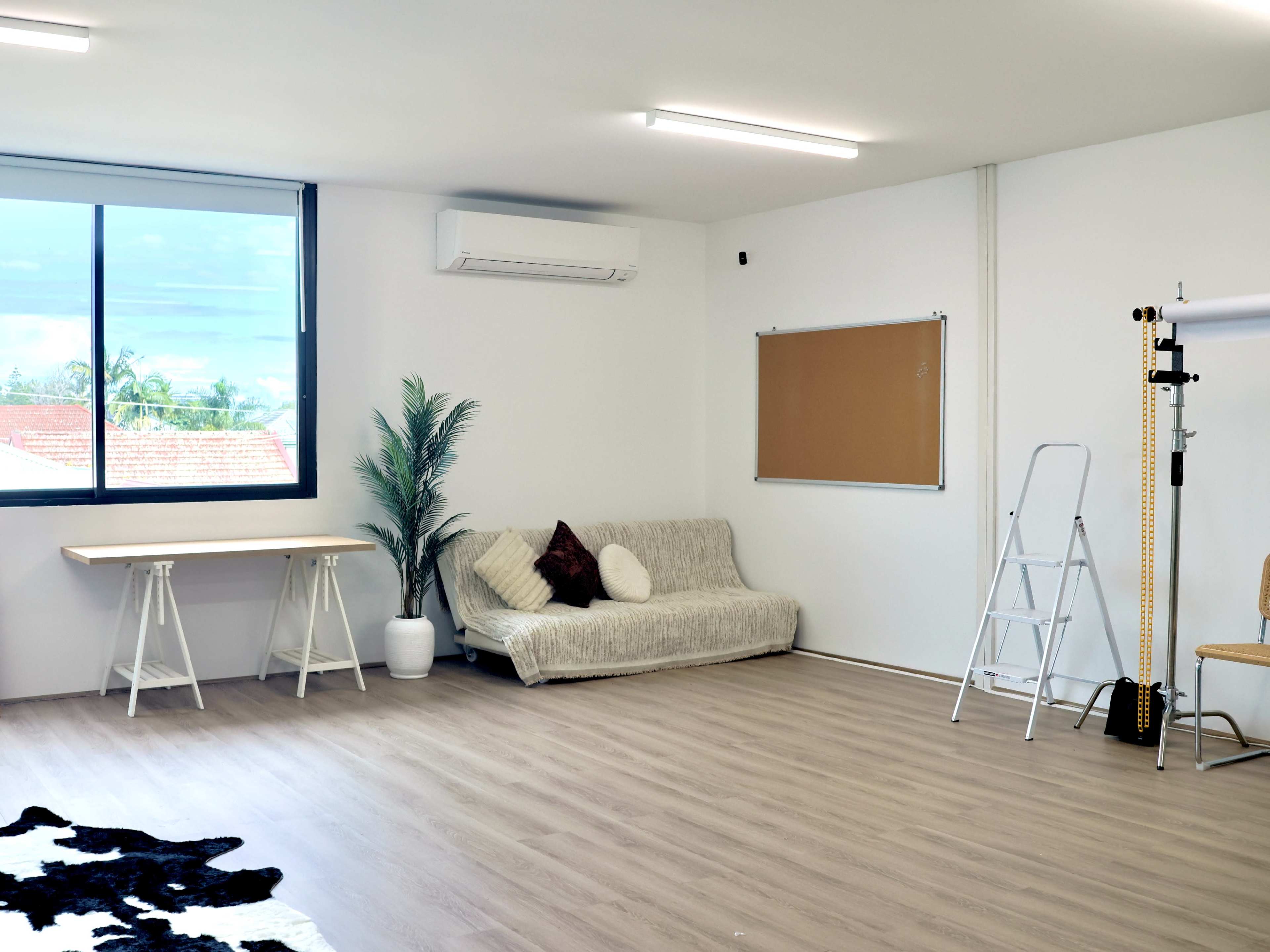 The image shows a bright room with a sofa, a desk, a ladder, and a bulletin board, featuring large windows that allow natural light to enter.