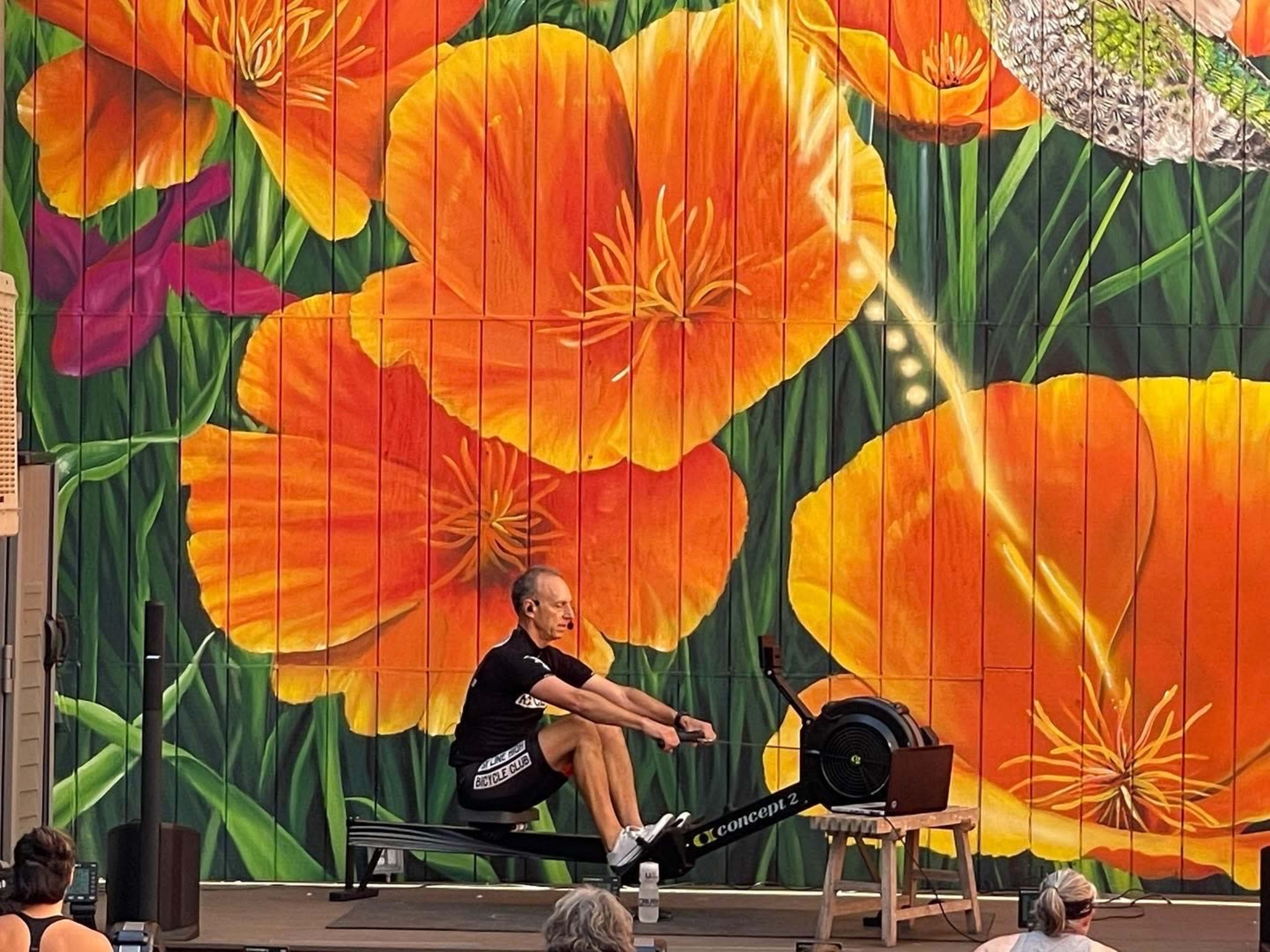 A person is rowing on a rowing machine in front of a colorful mural featuring large orange flowers.