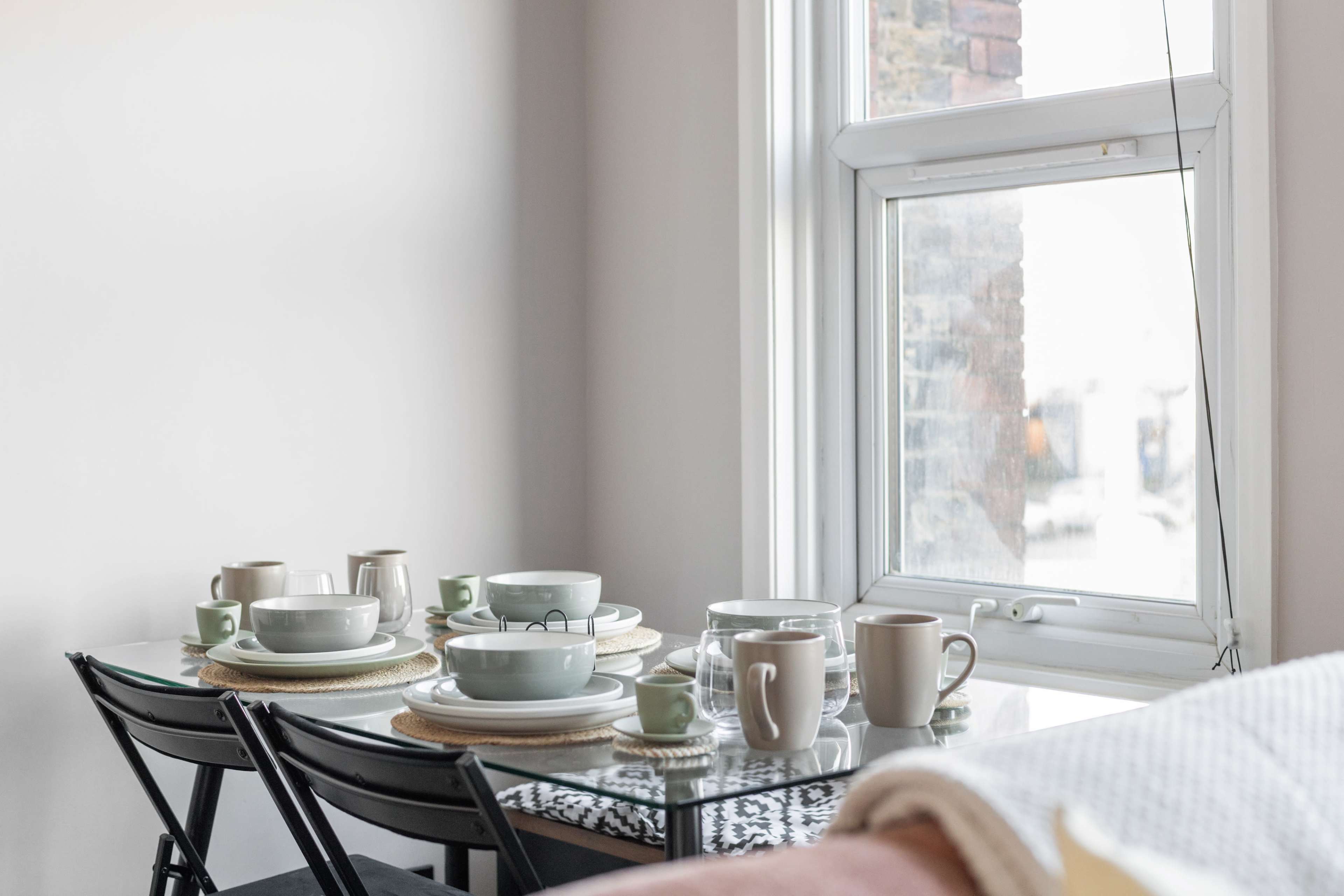 A dining table set with dinnerware and cups is positioned beside a window, allowing natural light to enter the room.