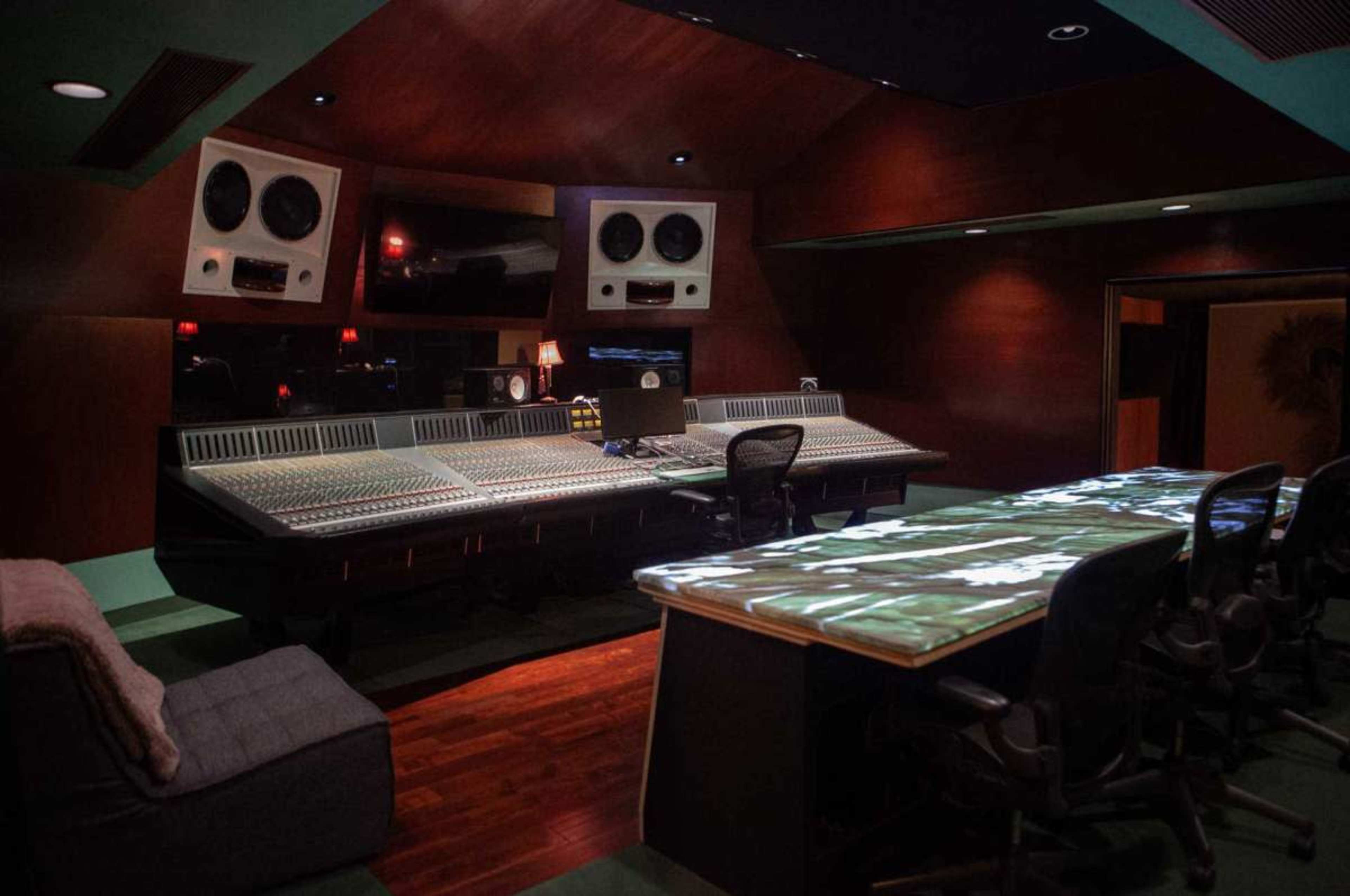 The image shows a professional music recording studio with a large mixing console, multiple speakers, and a spacious seating area.