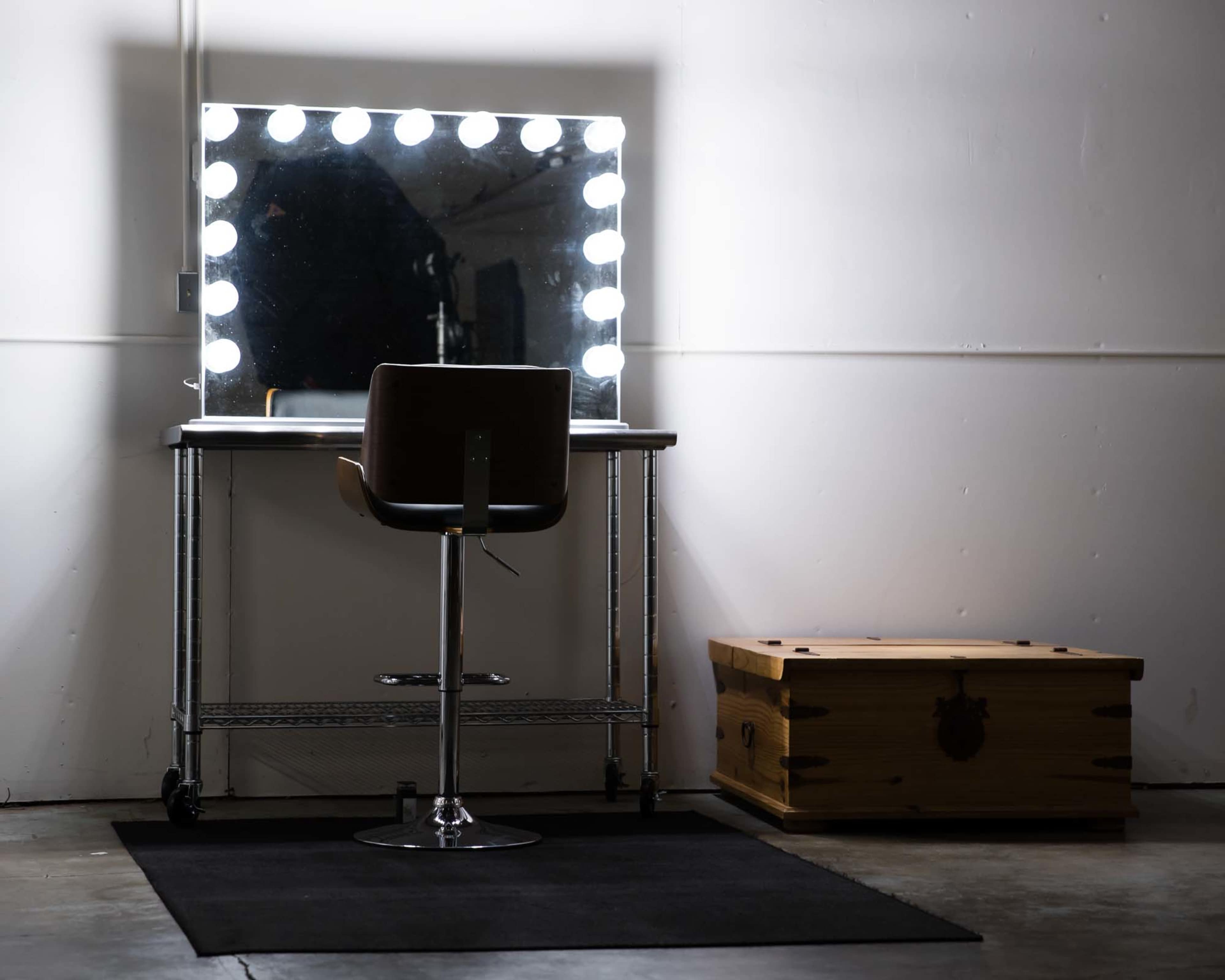 A lit makeup mirror is positioned above a rolling stool next to a wooden box on a black rug in a minimalist room.