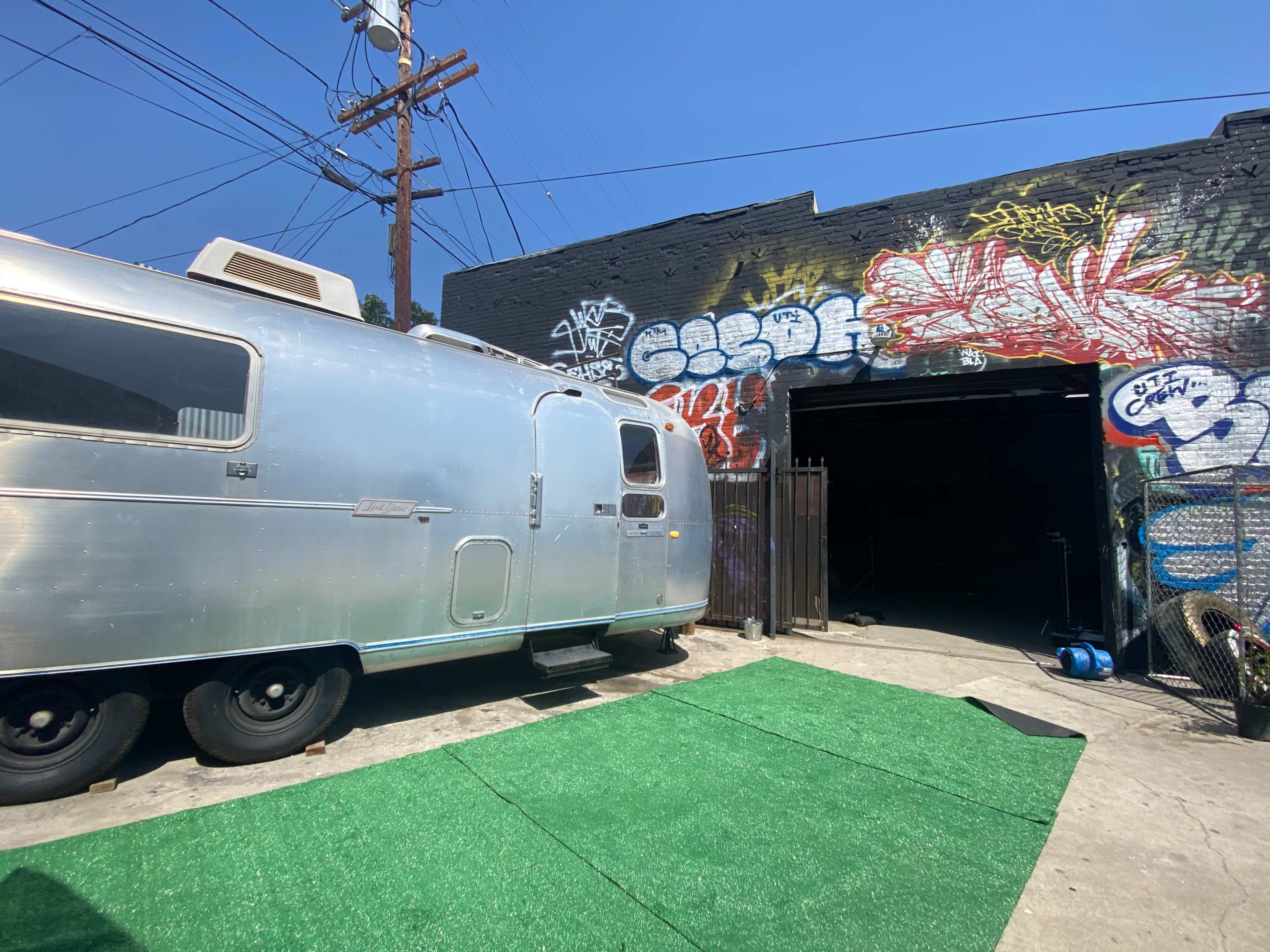 An Airstream trailer is parked next to a warehouse with graffiti-covered walls and a green turf area in front.