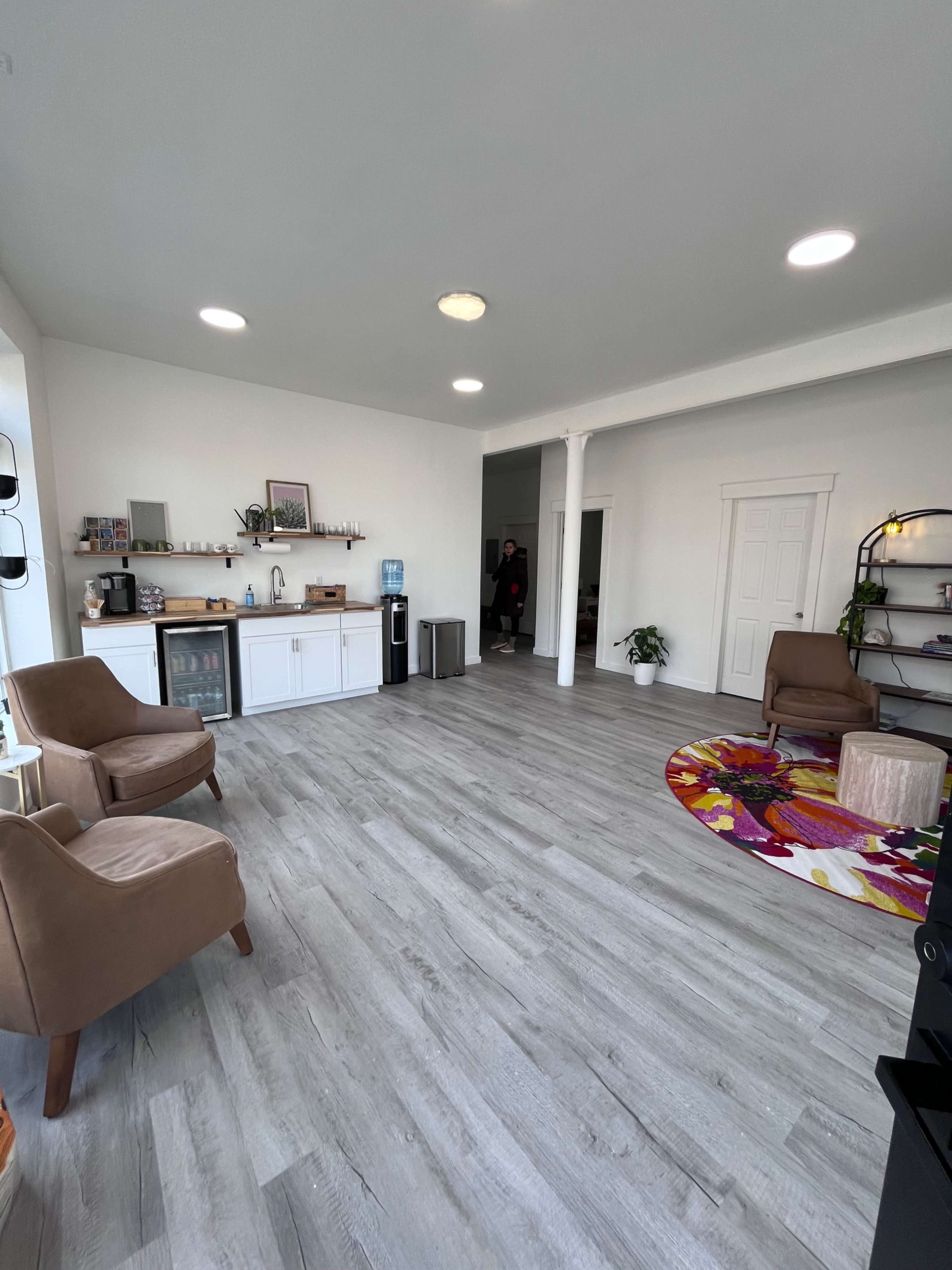 The image shows a bright and spacious room with a seating area, a kitchenette, and light-colored walls and flooring.