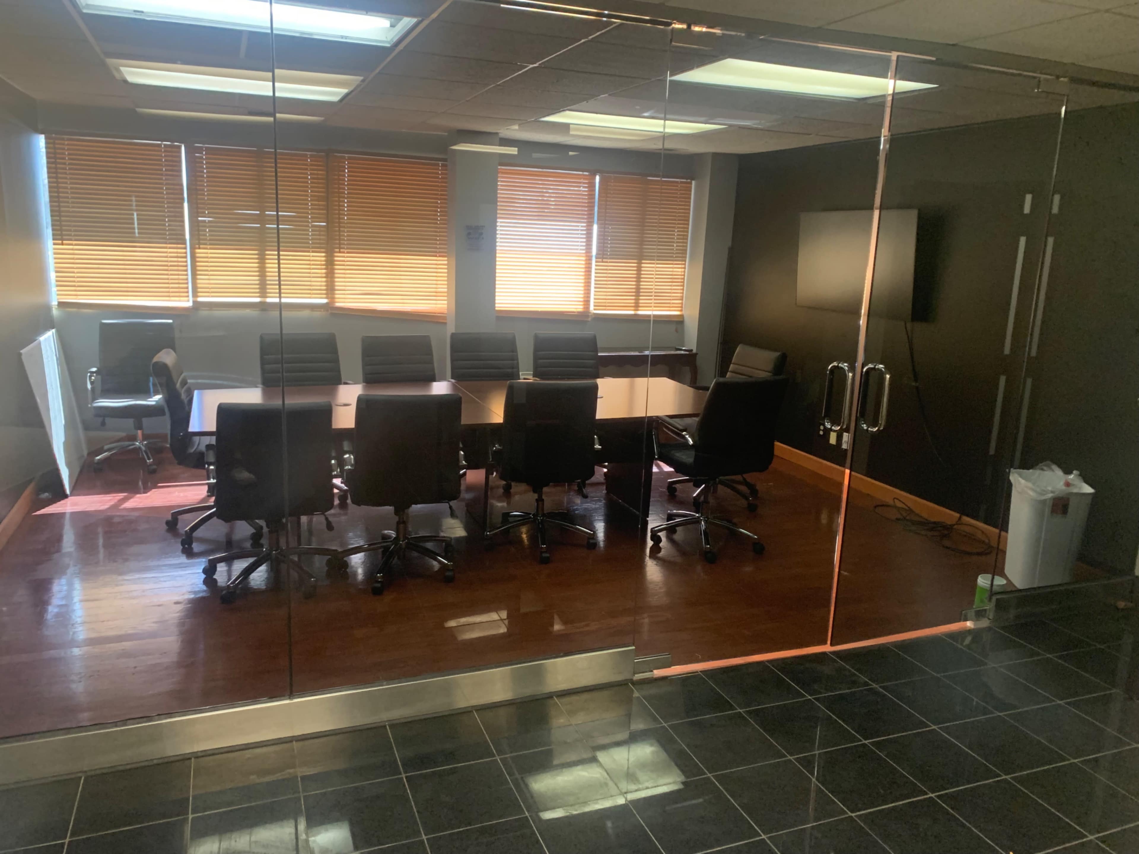 A glass-walled conference room features a long table surrounded by black chairs and large windows with wooden blinds.