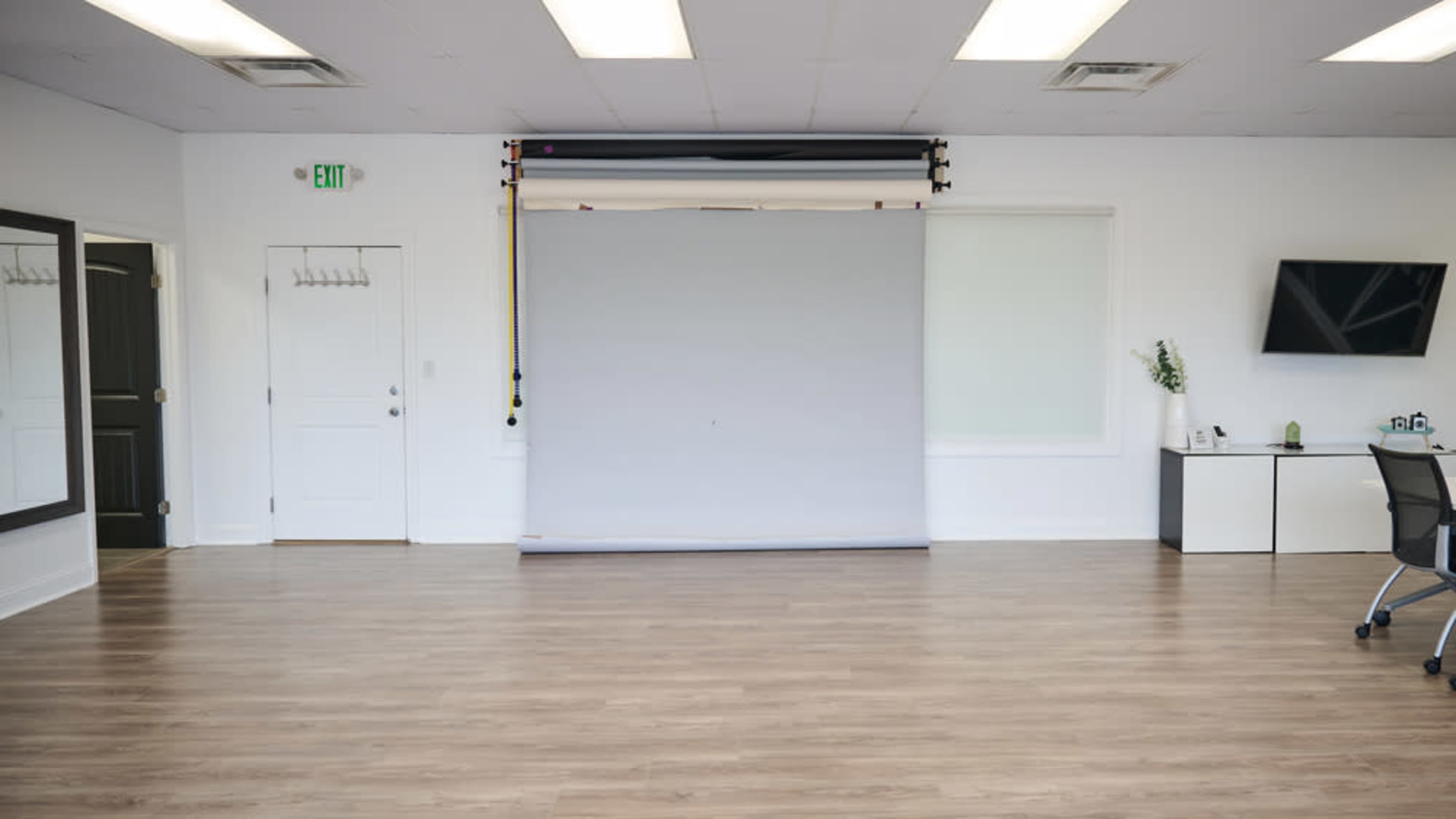 The image shows a spacious indoor room featuring a blank gray backdrop, a desk with a chair, a wall-mounted television, and a large mirror.