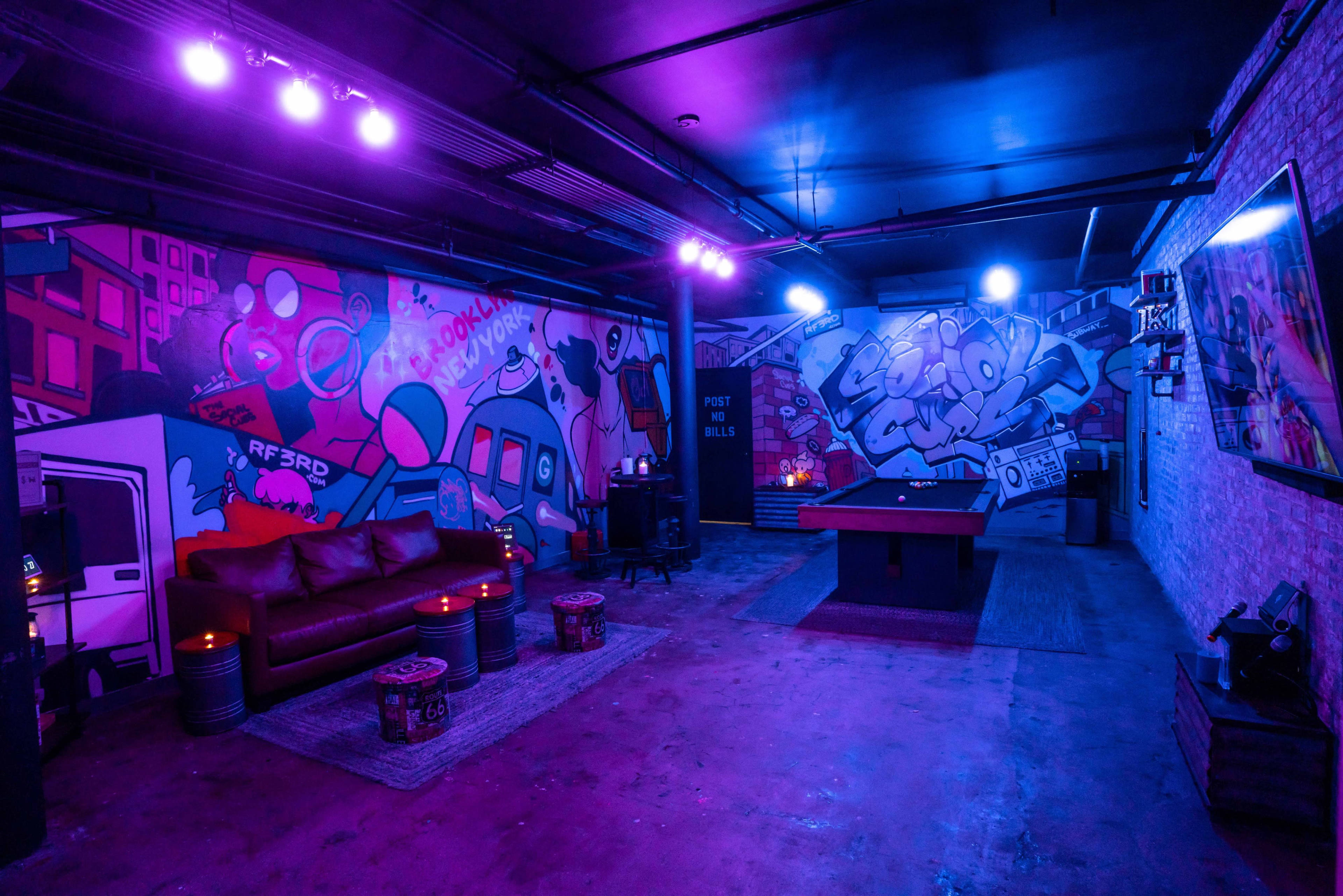 The image shows a dimly lit entertainment space featuring a pool table, colorful graffiti murals on the walls, and a couch with ambient lighting.