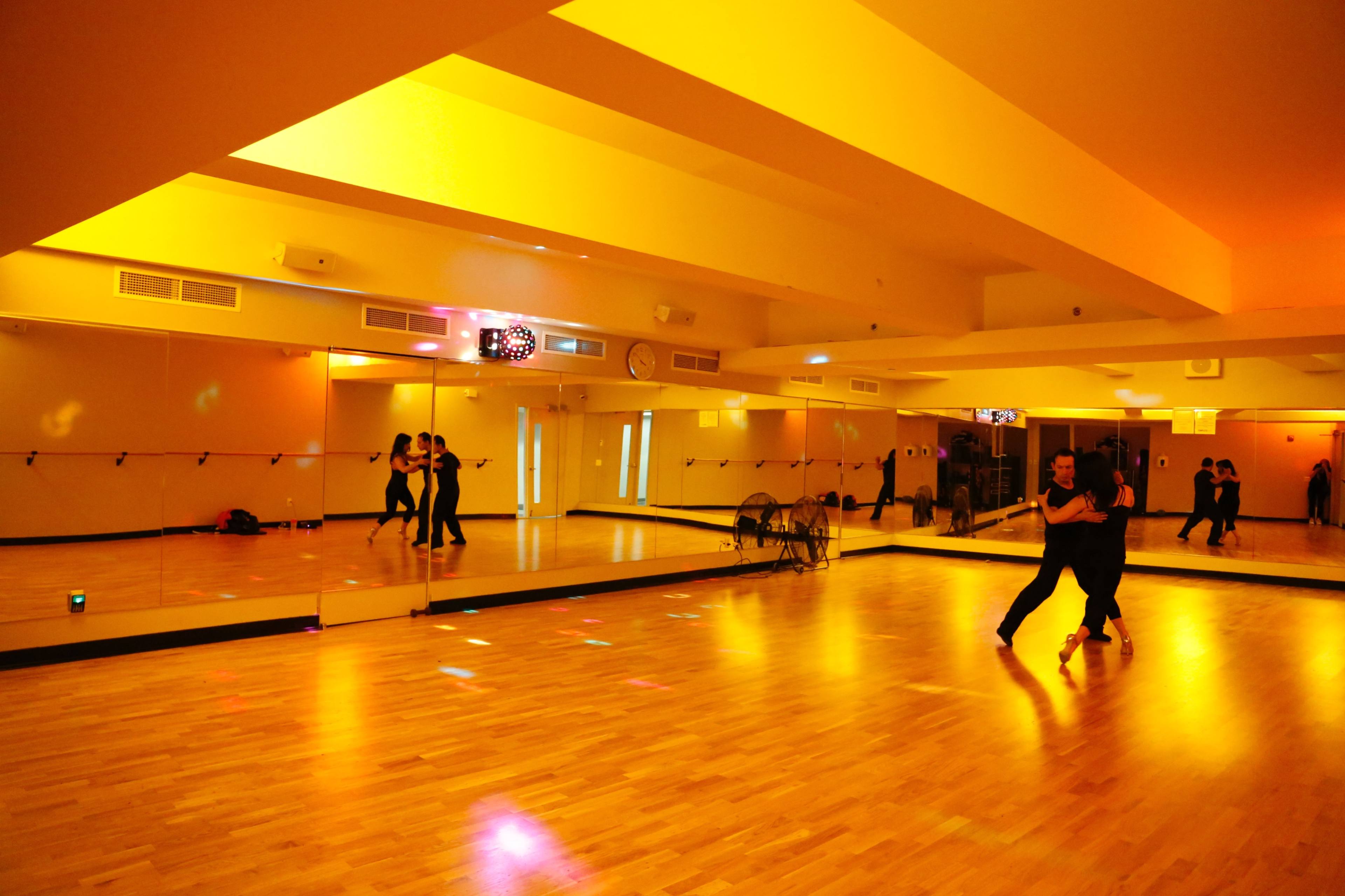 A couple dances in a brightly lit studio with mirrors and a wooden floor.