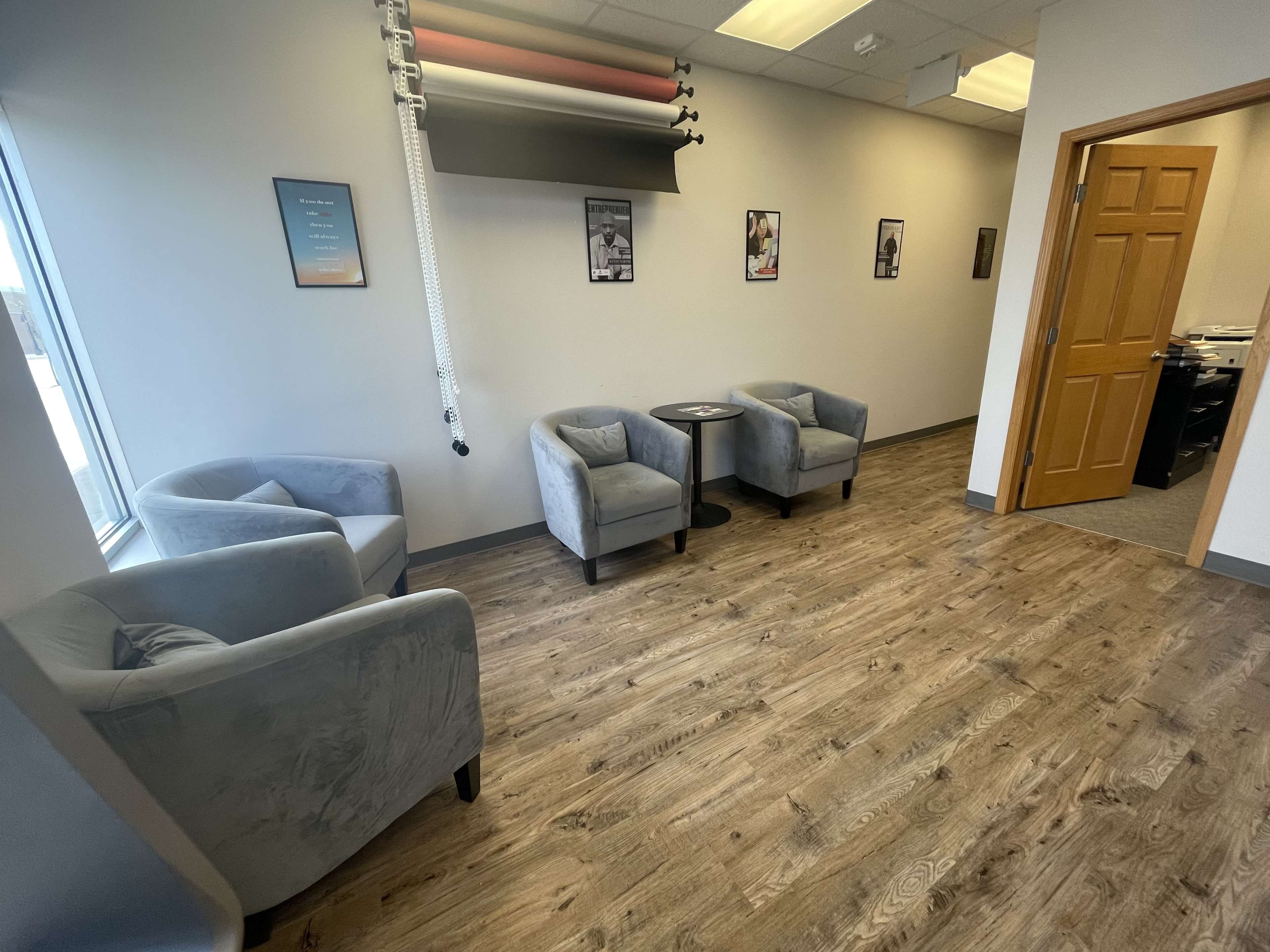 A small waiting area with four gray armchairs and a side table, located near a window in a room with light-colored walls and wooden flooring.