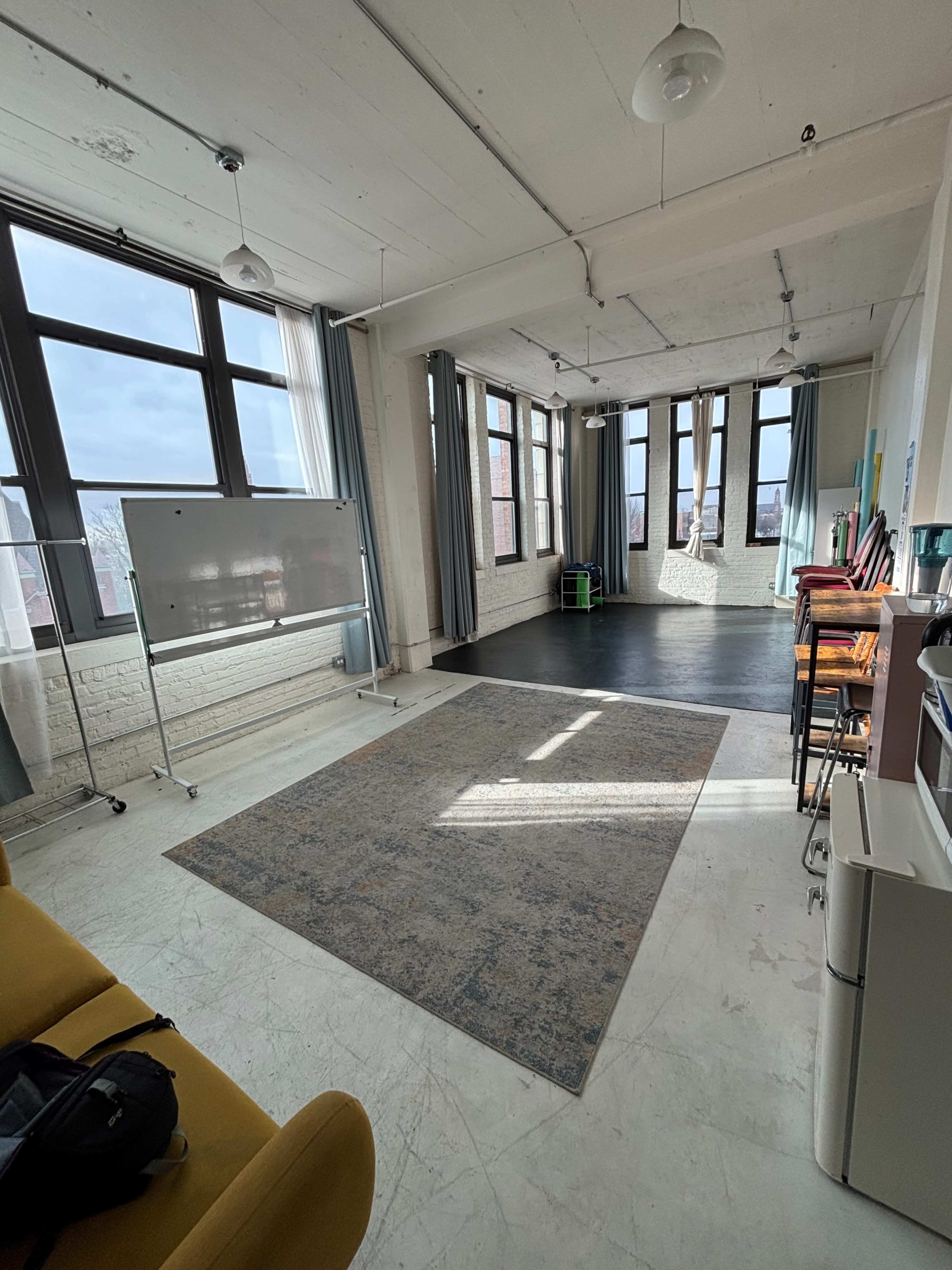 The image shows a spacious, well-lit room with large windows, a gray rug on the floor, and seating along the wall.