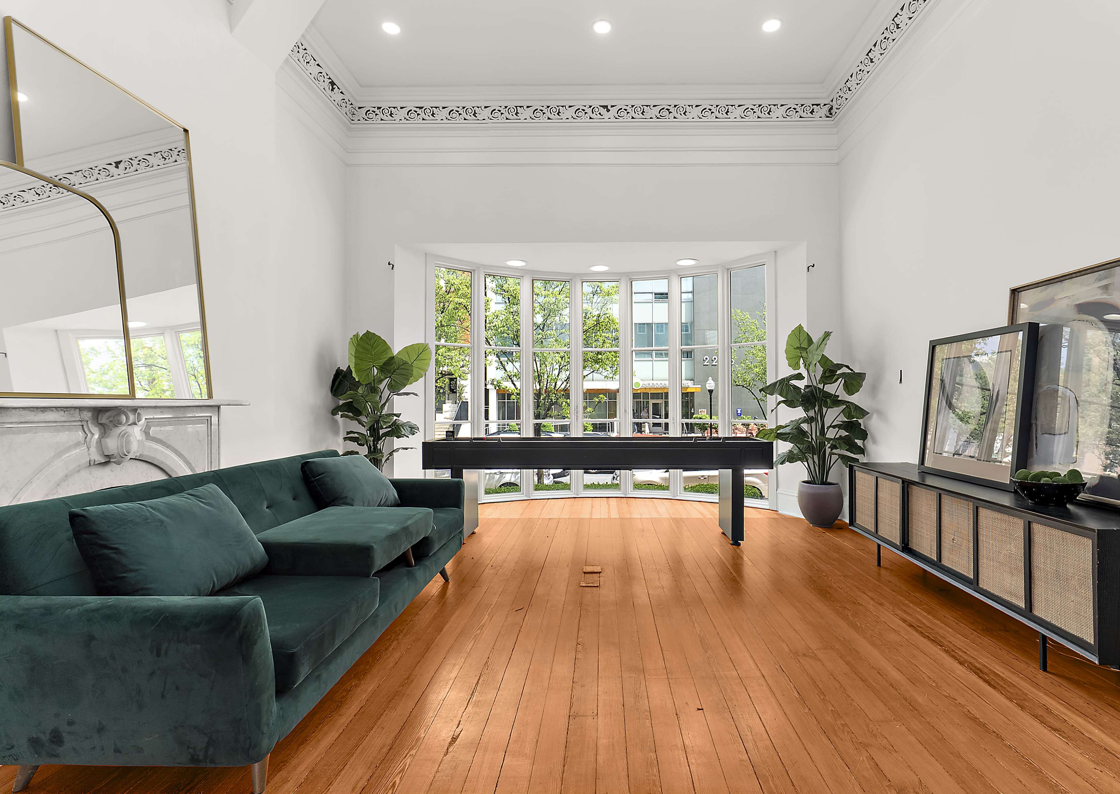 The scene depicts a modern living room with a green sofa, a long table, and large windows facing a tree-lined street.