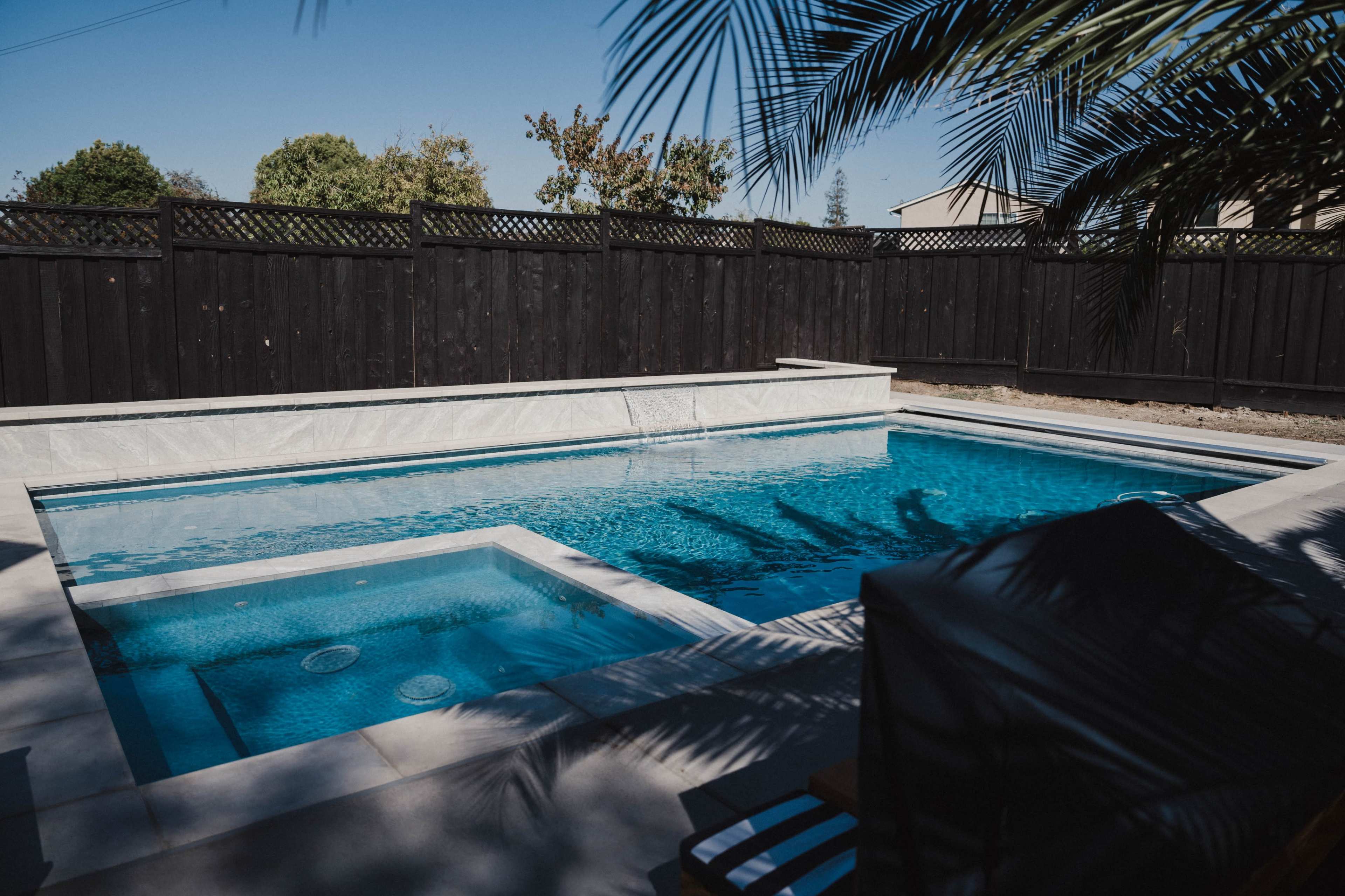 A clear swimming pool surrounded by a black wooden fence and palm trees.