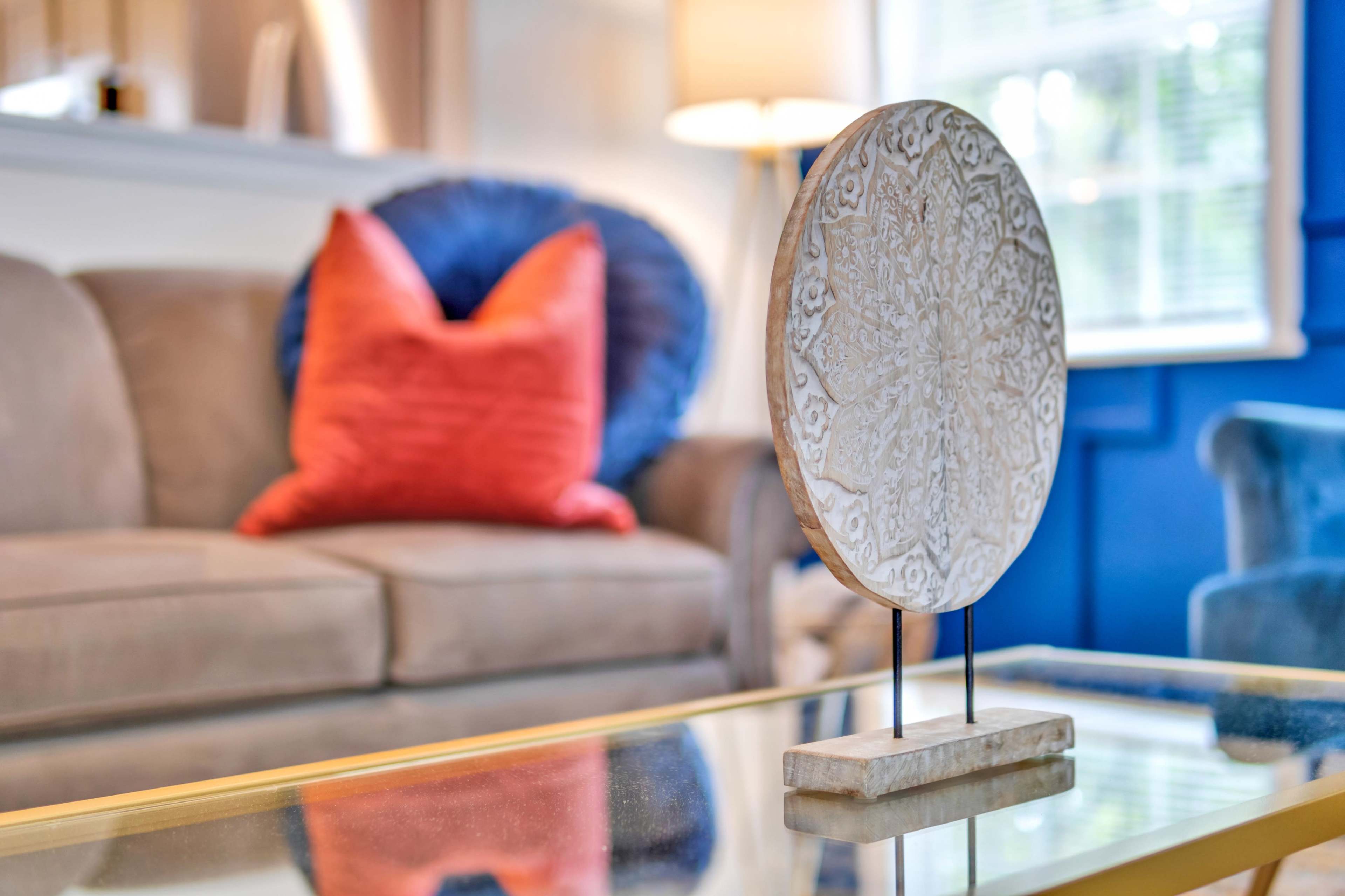 A decorative circular object is displayed on a glass coffee table in a living room featuring a beige couch and a blue wall.