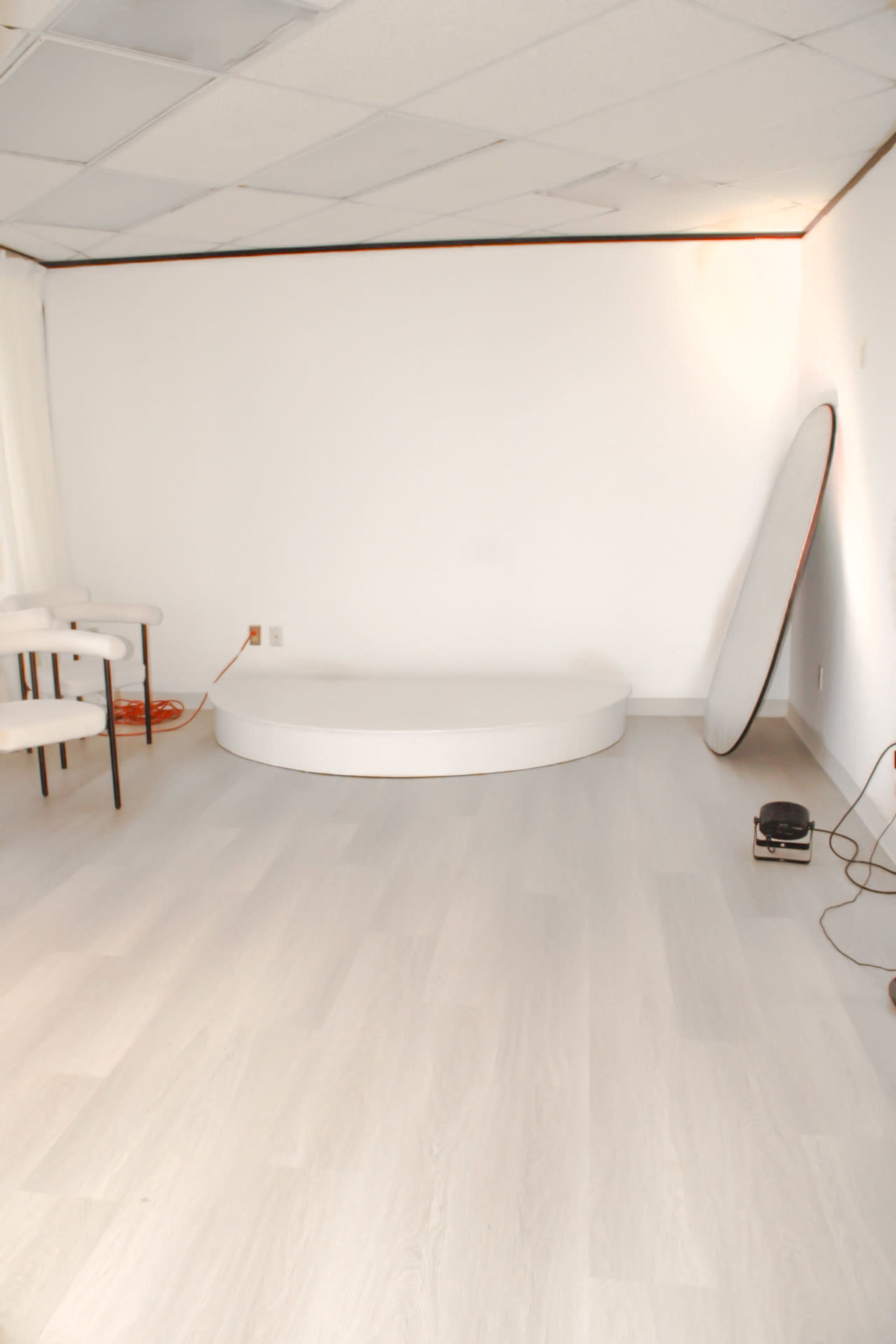 The image shows a minimalistic, empty room with light-colored wooden flooring, a small circular stage, and a full-length mirror leaning against the wall.
