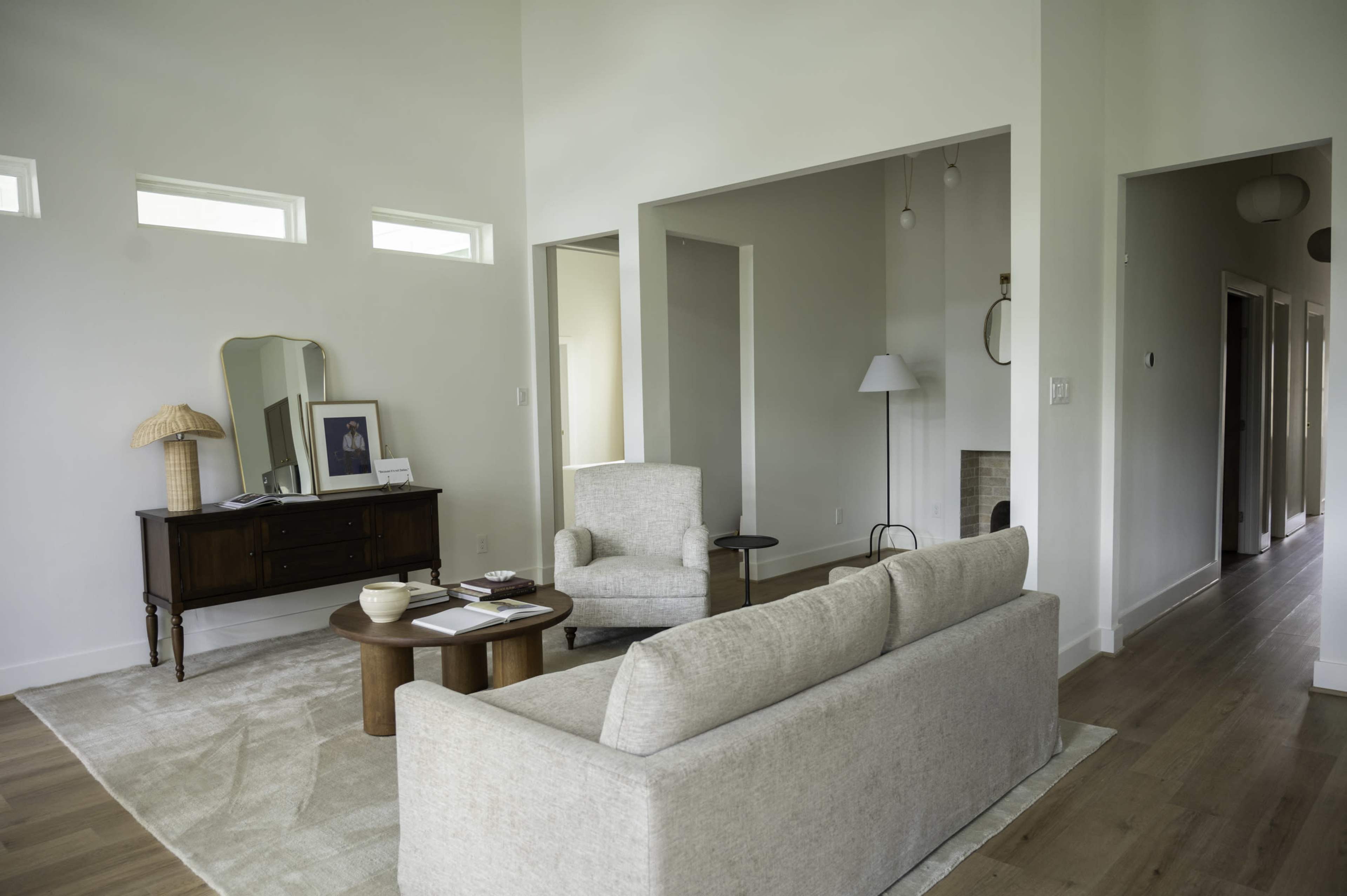 The image depicts a modern, minimalistic living room with a light-colored sofa, a round wooden coffee table, and a wooden sideboard featuring a framed photo.