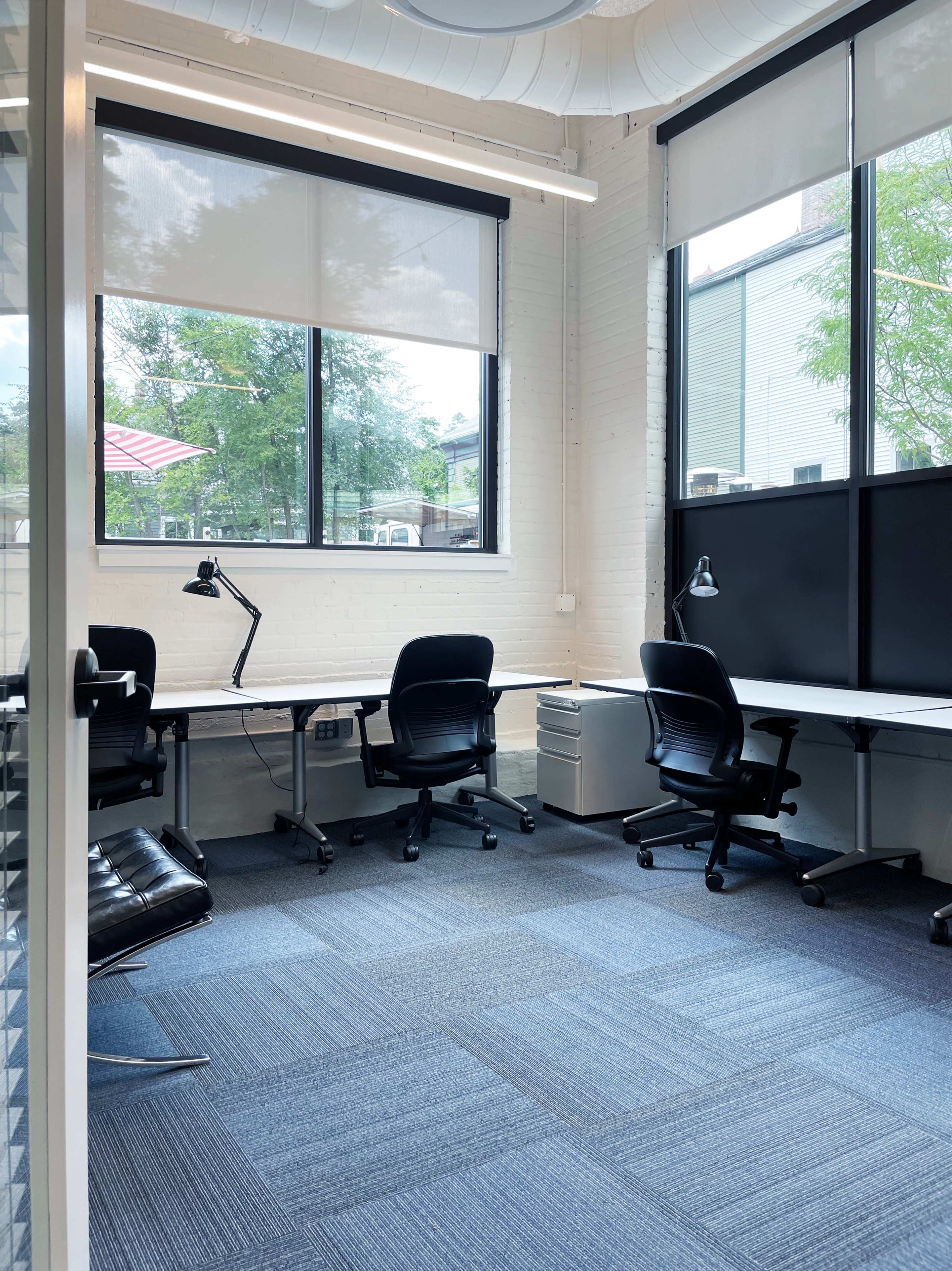 A bright office space with two desks, ergonomic chairs, and large windows allowing natural light to enter.