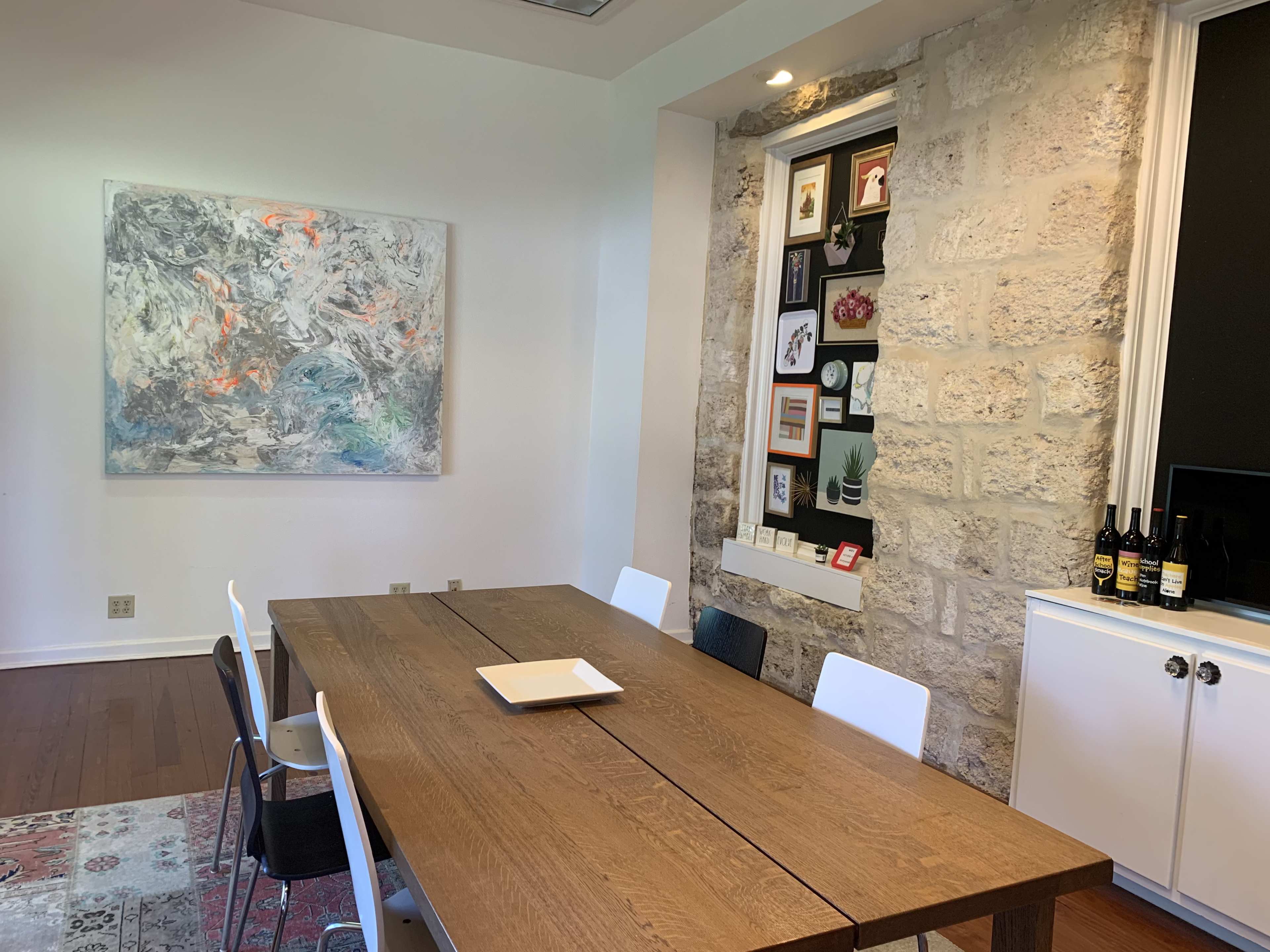 A modern dining area featuring a wooden table surrounded by white and black chairs, with an abstract painting on the wall and a stone nook displaying framed pictures and decor.
