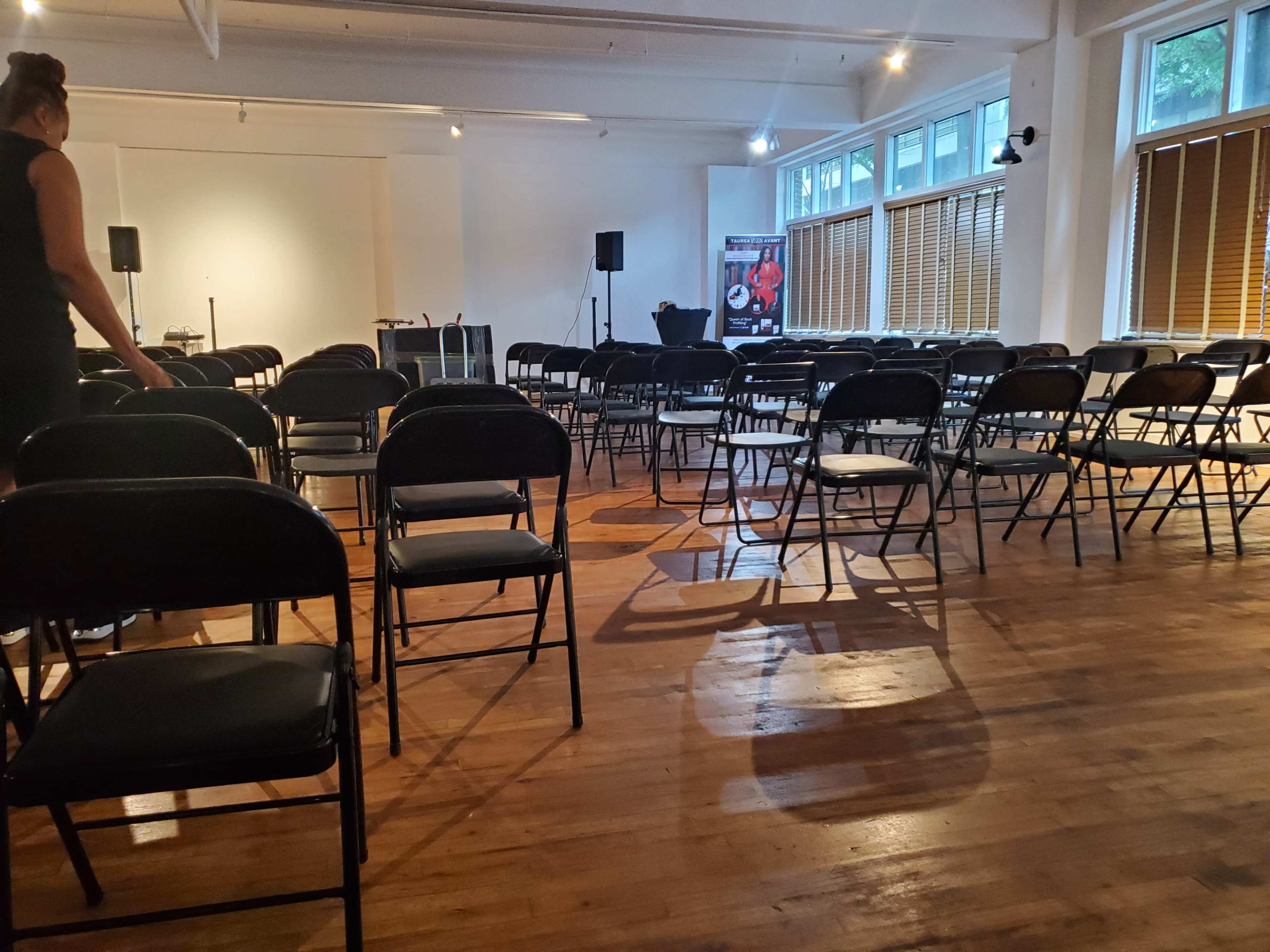 A spacious room with rows of black folding chairs arranged on a wooden floor, and a person walking towards the front where equipment is set up.