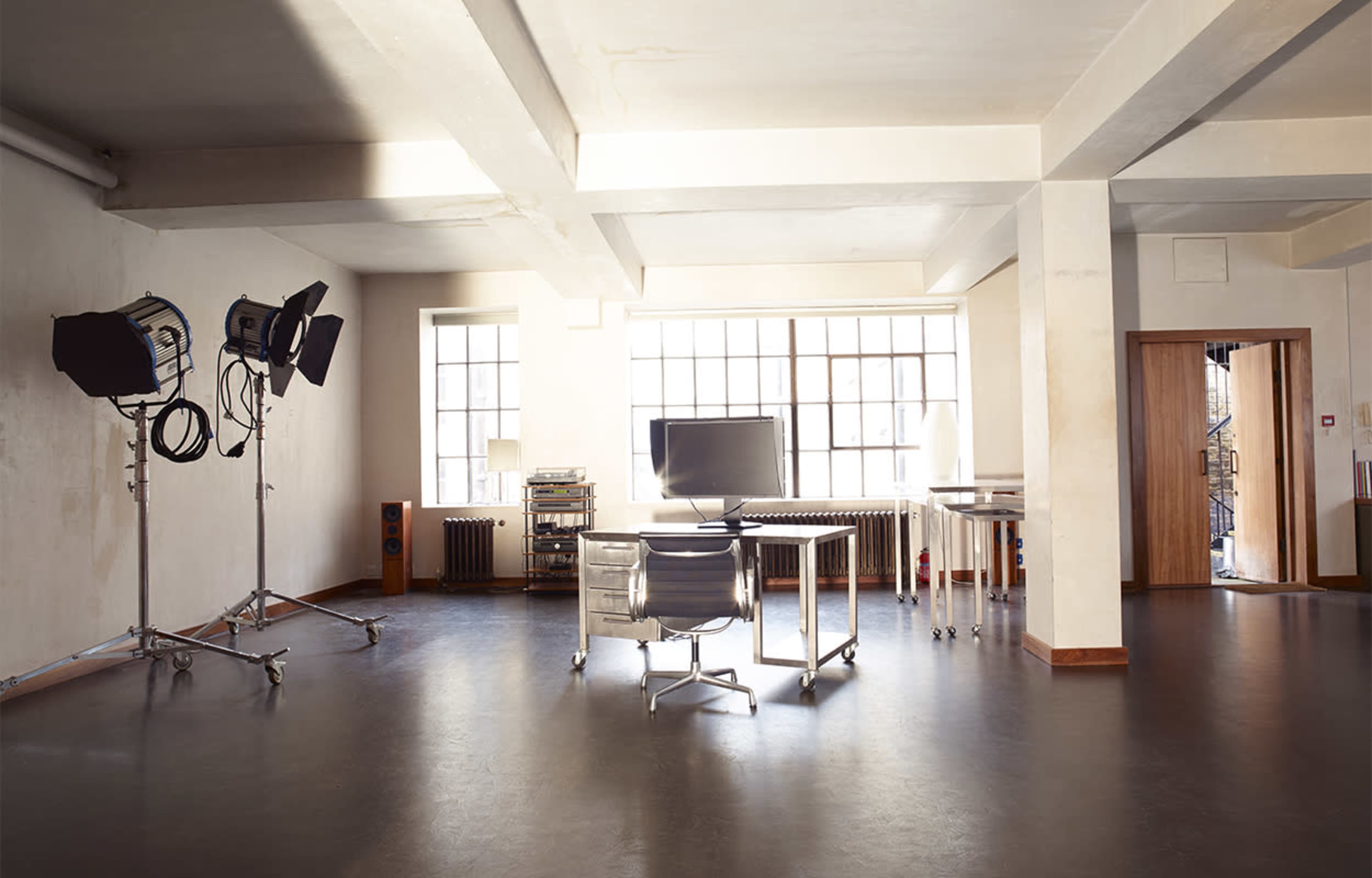 A spacious, minimalist studio with a desk, a computer monitor, and multiple lighting setups beside large windows.