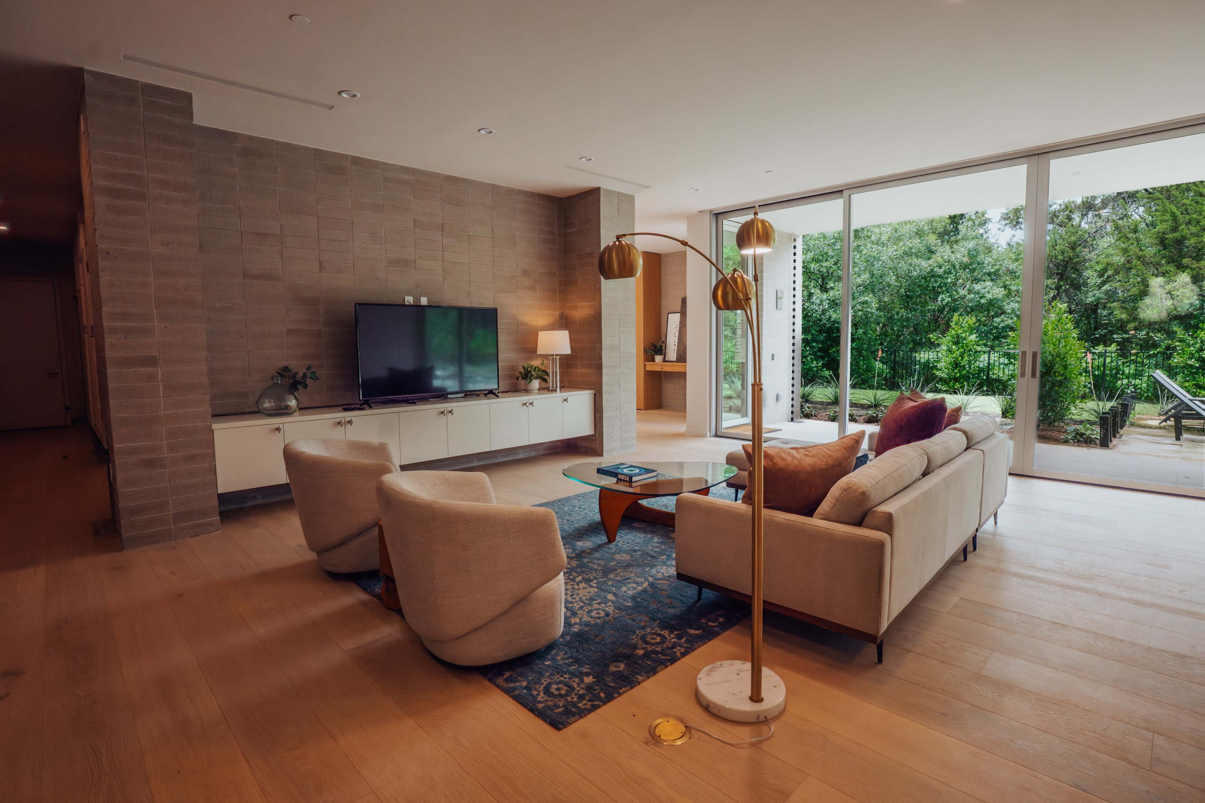 The scene shows a modern living room with a light-colored sofa, two armchairs, a television on a console, and large glass doors that open to a green outdoor space.