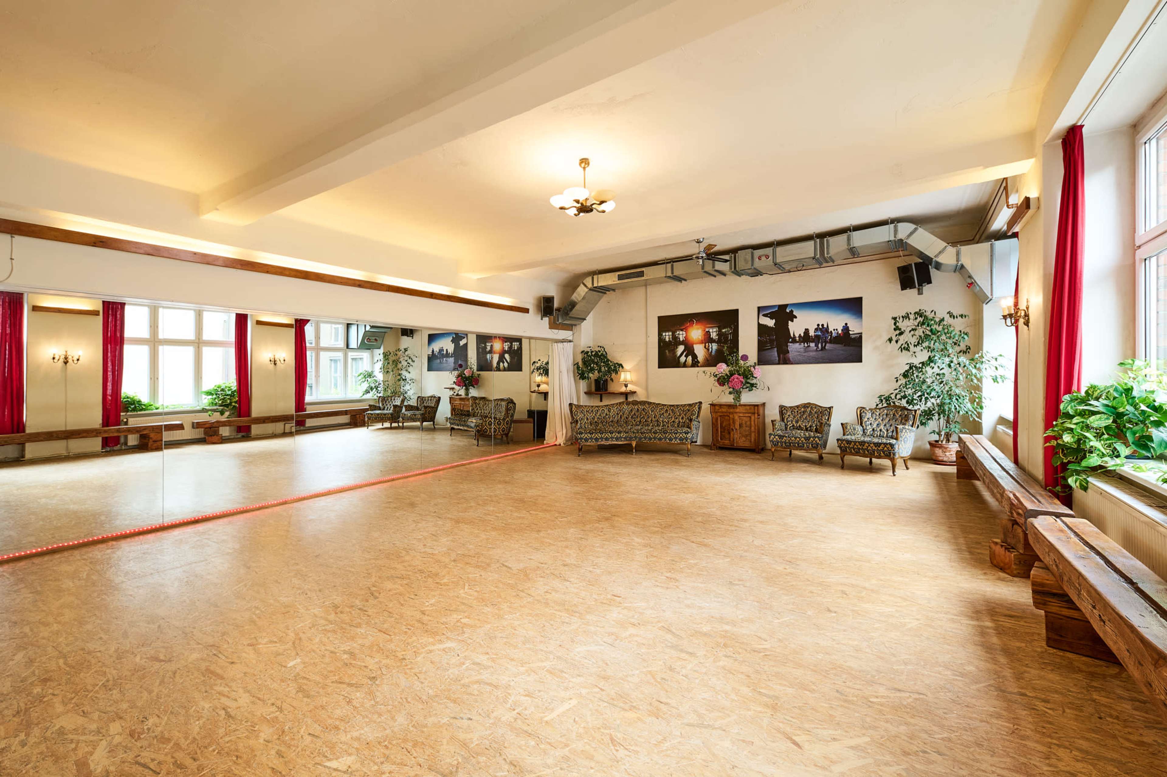 A spacious dance studio features a mirrored wall, wooden flooring, and decorative plants, with seating along the edges and large windows letting in natural light.