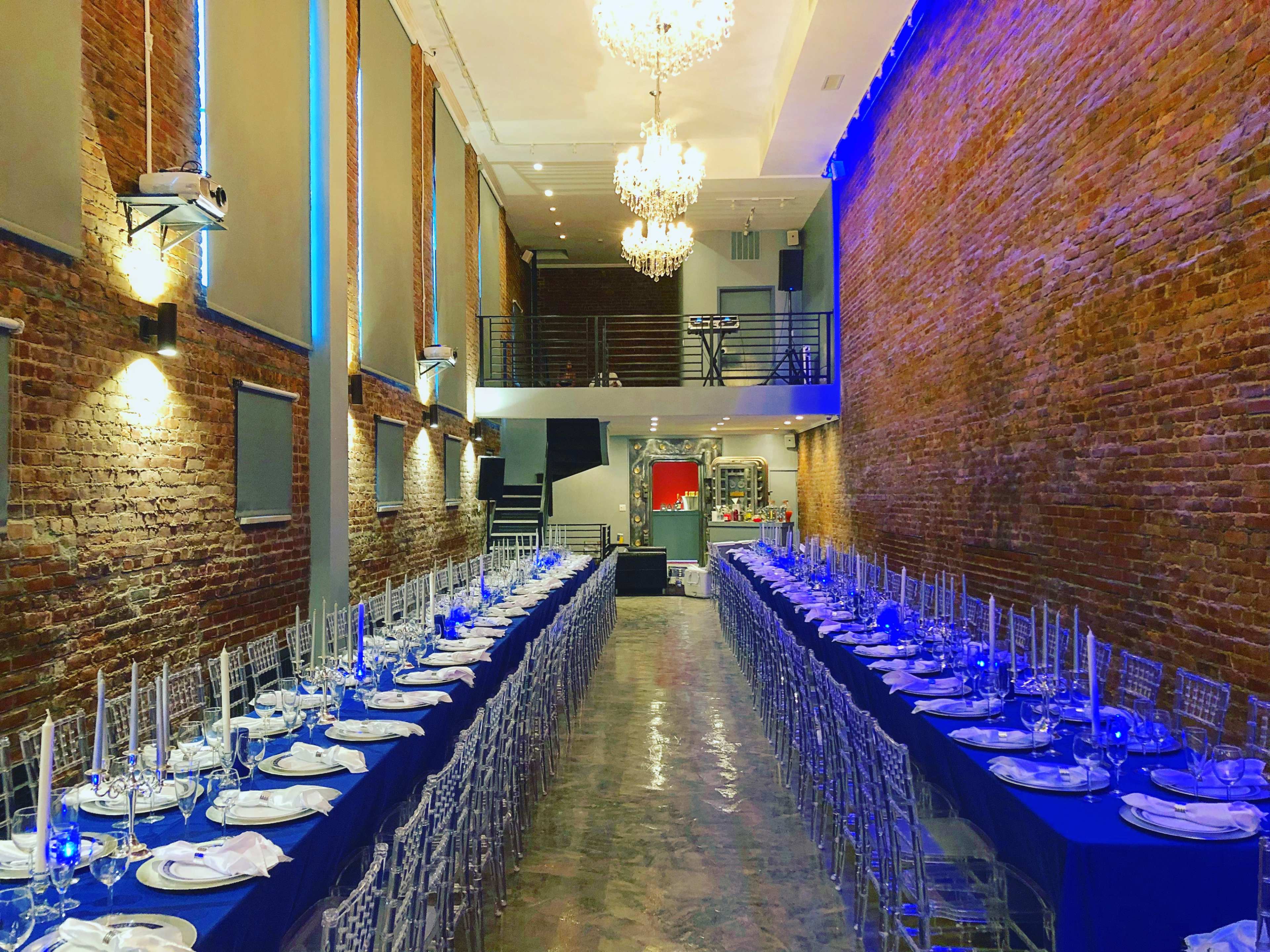 A long dining table set for an event in a brick-walled hall, illuminated by chandeliers and blue lighting.