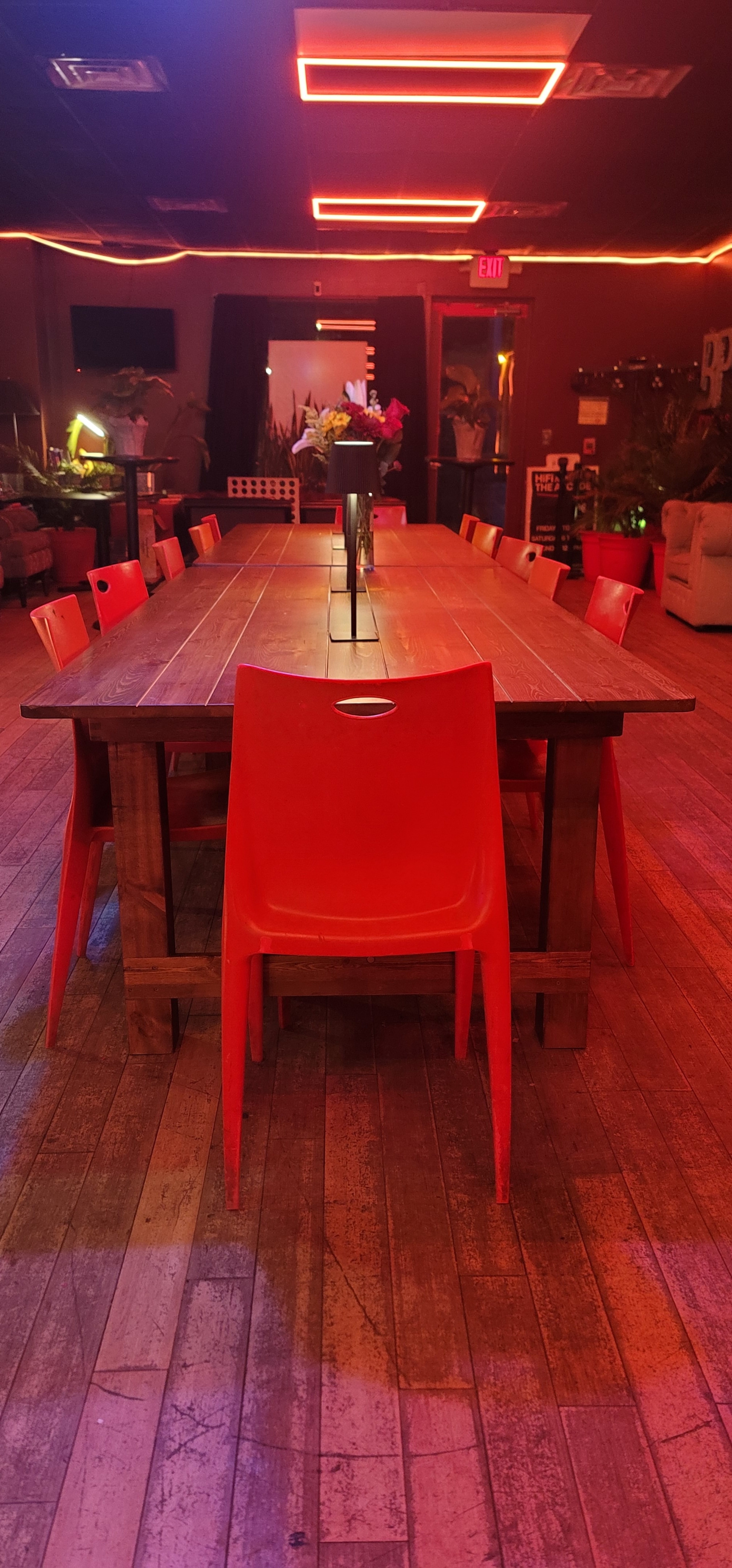 A large wooden table surrounded by red plastic chairs is illuminated by vibrant neon lights in a dimly lit room.