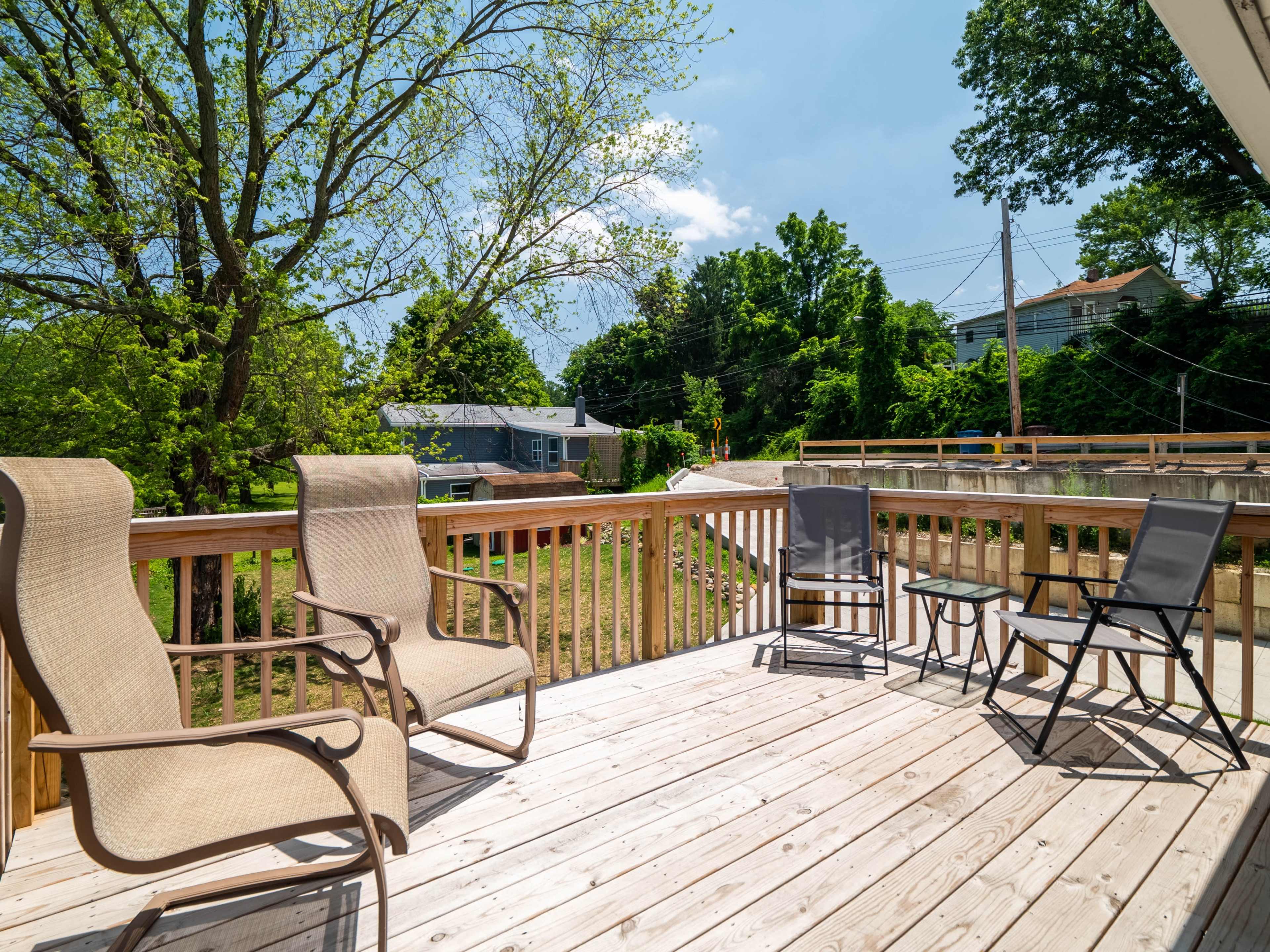 A wooden deck furnished with four chairs overlooking a green, tree-lined area and a road.