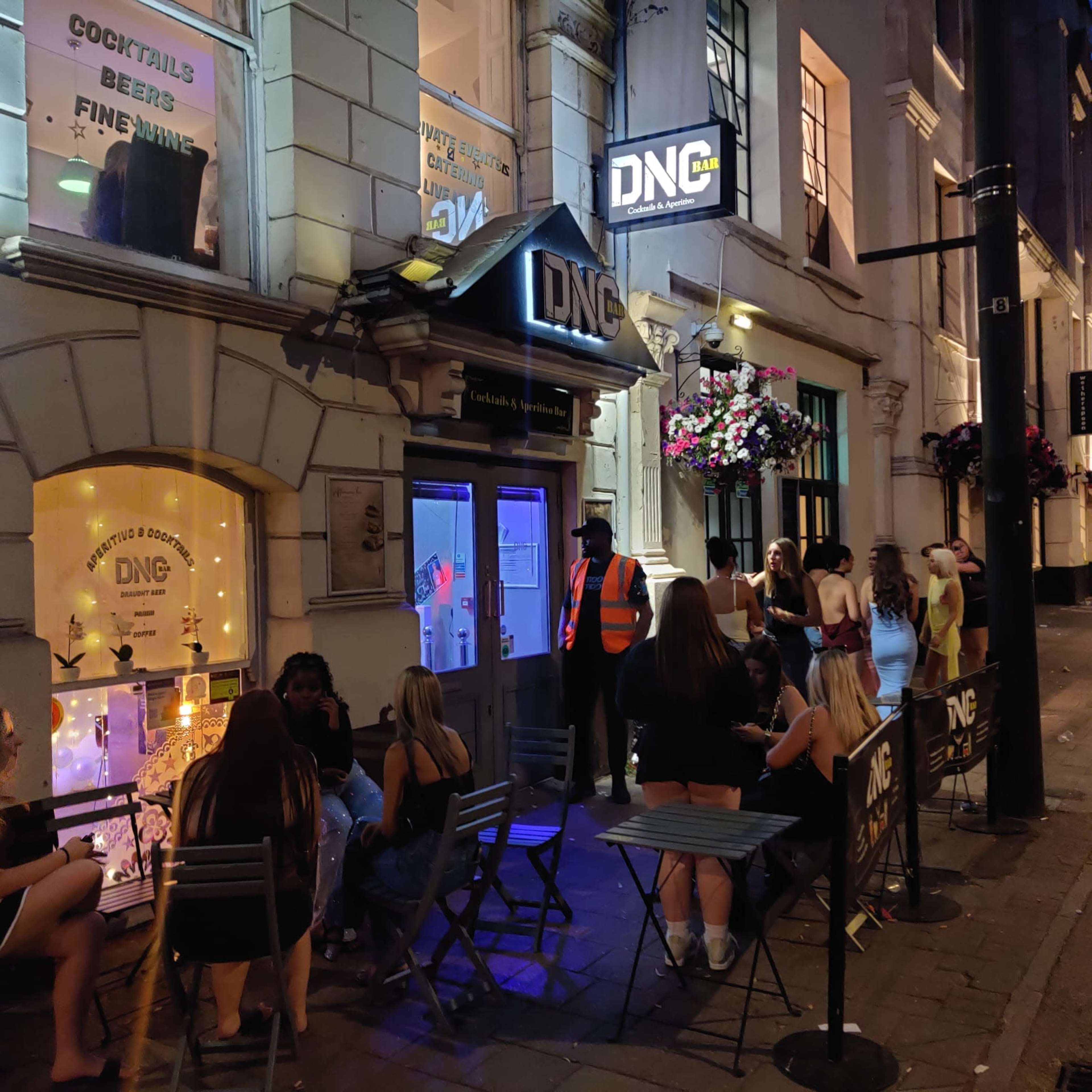 The image shows a bar entrance labeled "DNG" with patrons sitting outside on a street lined with flowers.