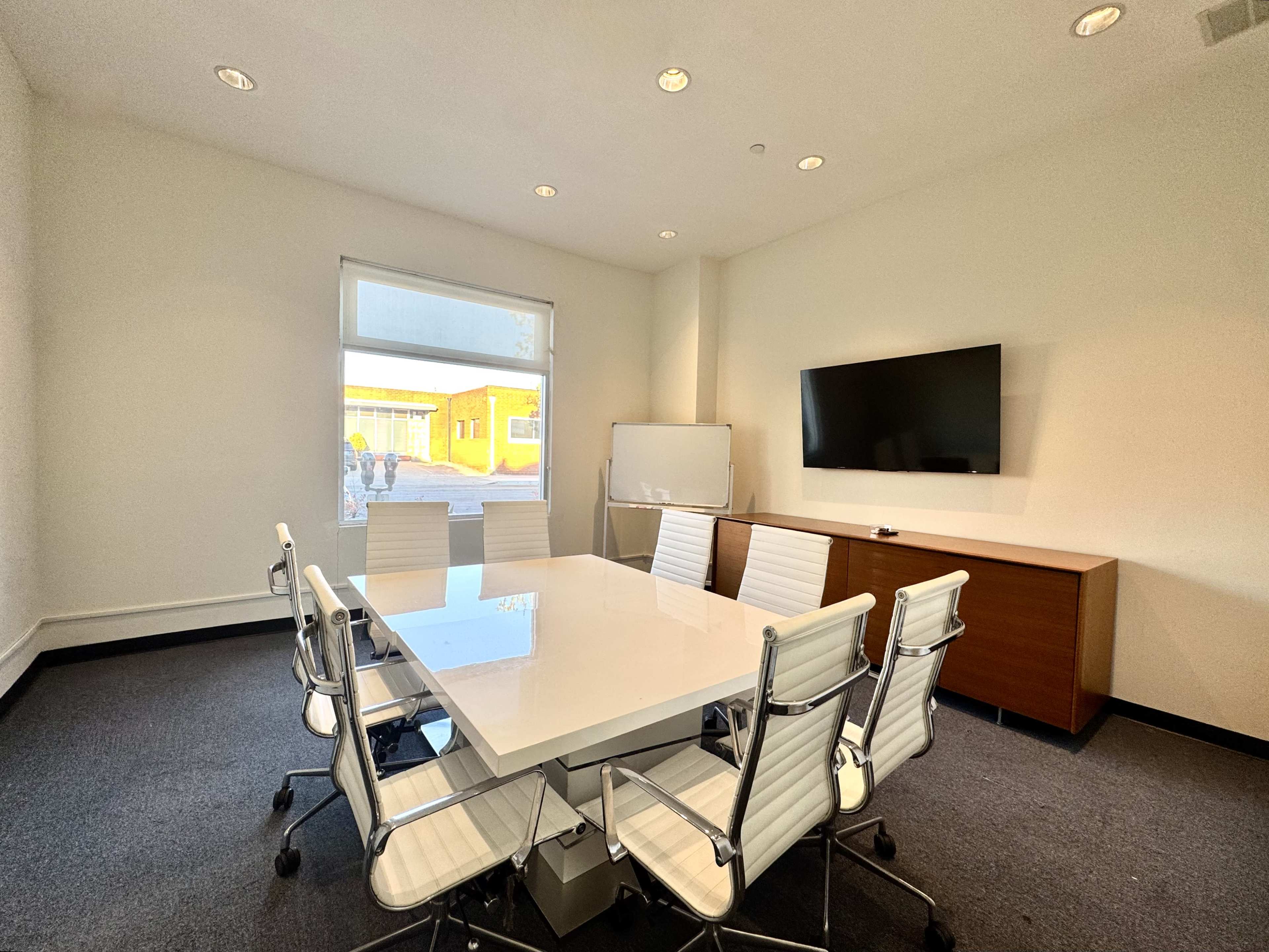 A modern conference room features a large white table surrounded by eight ergonomic chairs, a wall-mounted television, and a whiteboard.