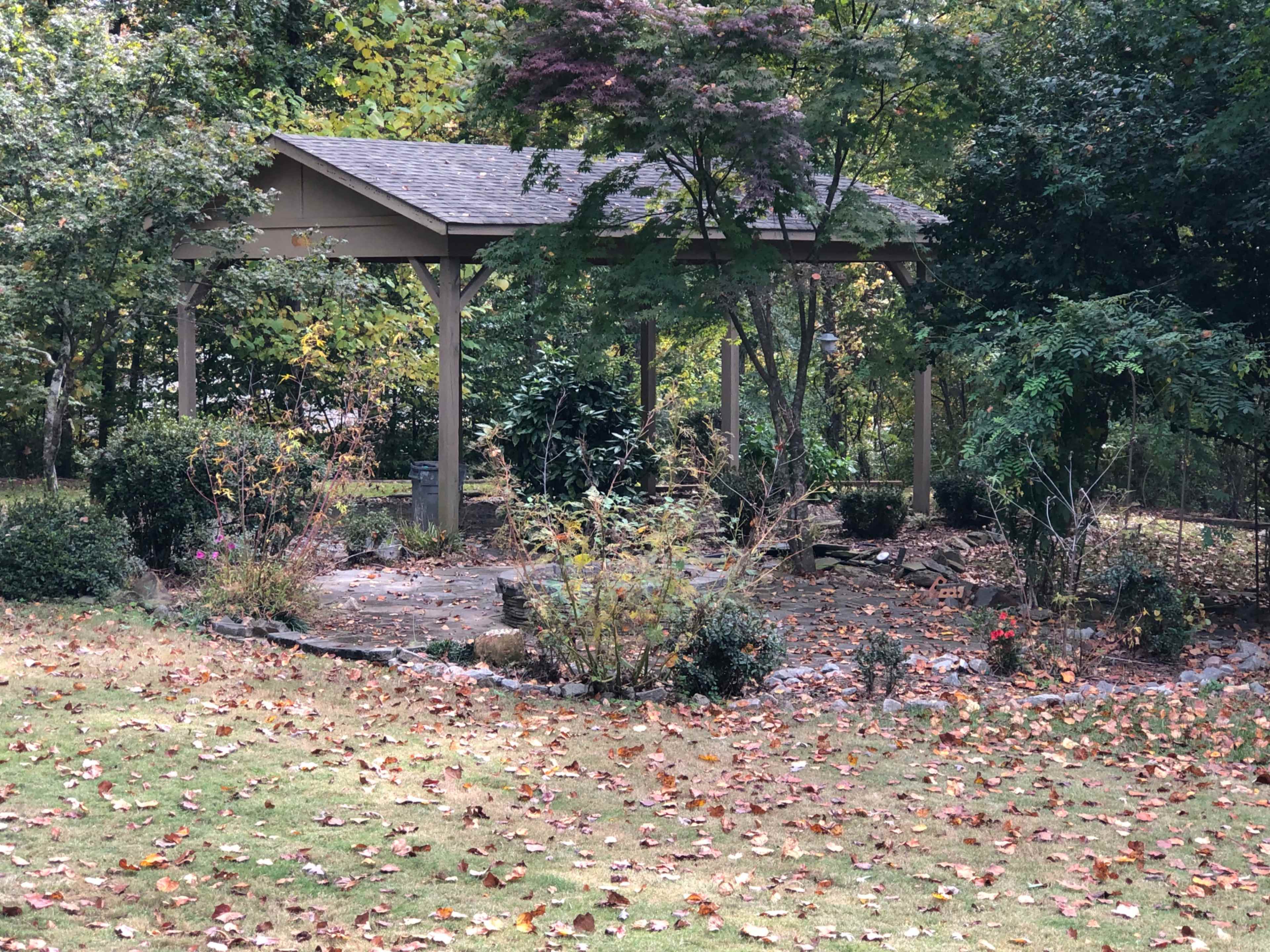 A small pavilion is surrounded by shrubbery and fallen leaves in a grassy area.