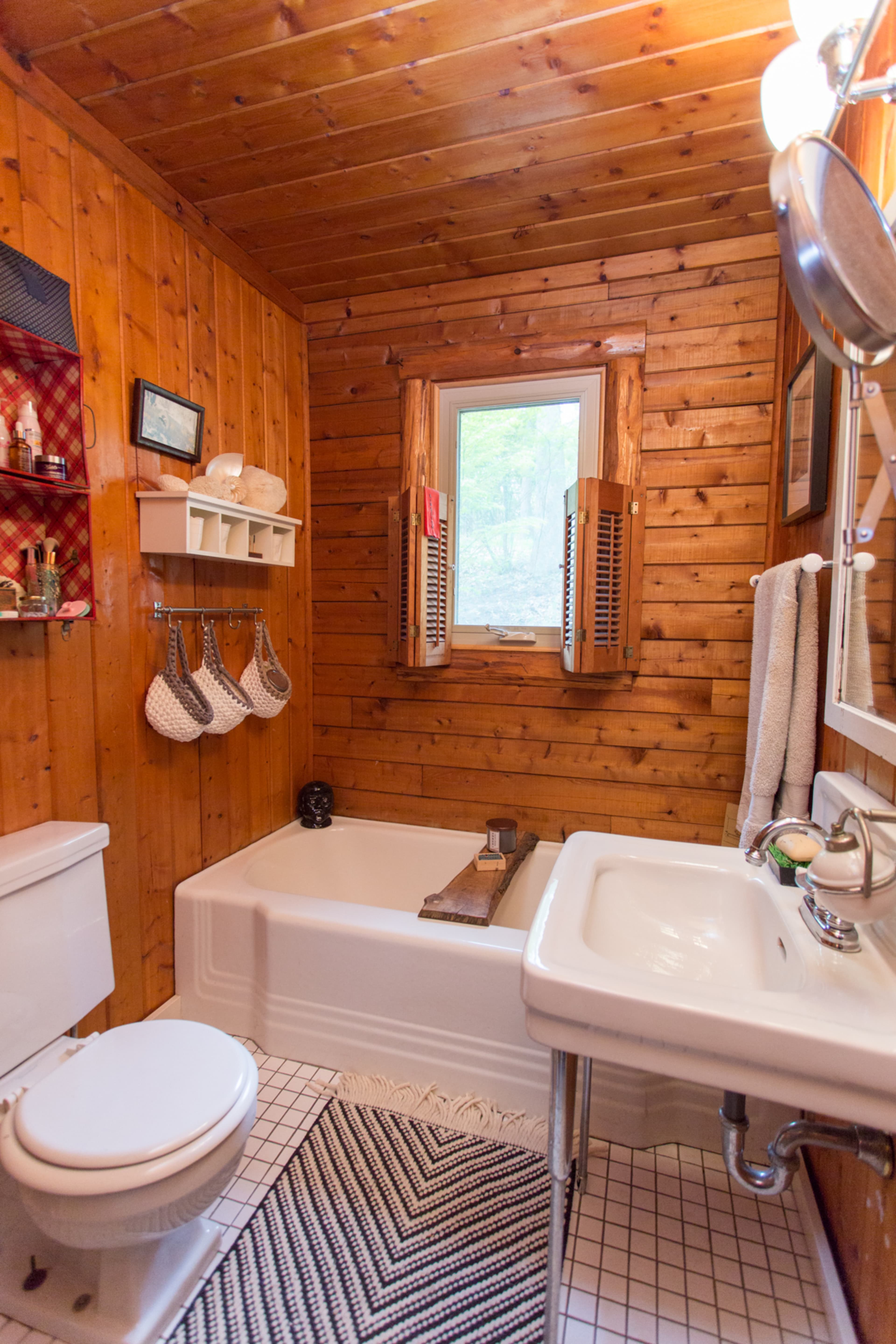 A small, rustic bathroom with wooden walls, a bathtub, a sink, a window, and organized wall storage.