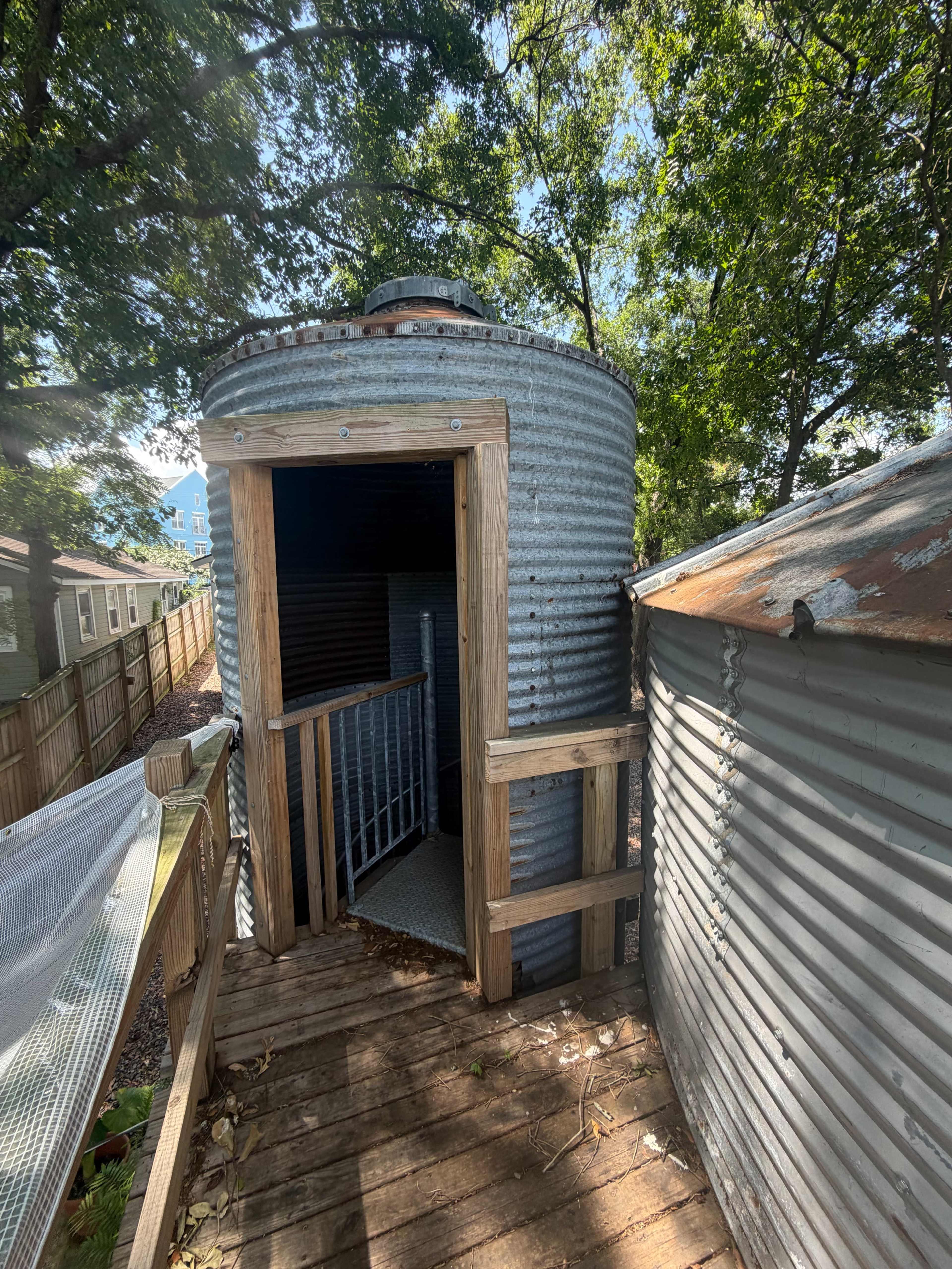 The image shows a circular, corrugated metal structure with a wooden entrance and a deck, surrounded by trees and other buildings.