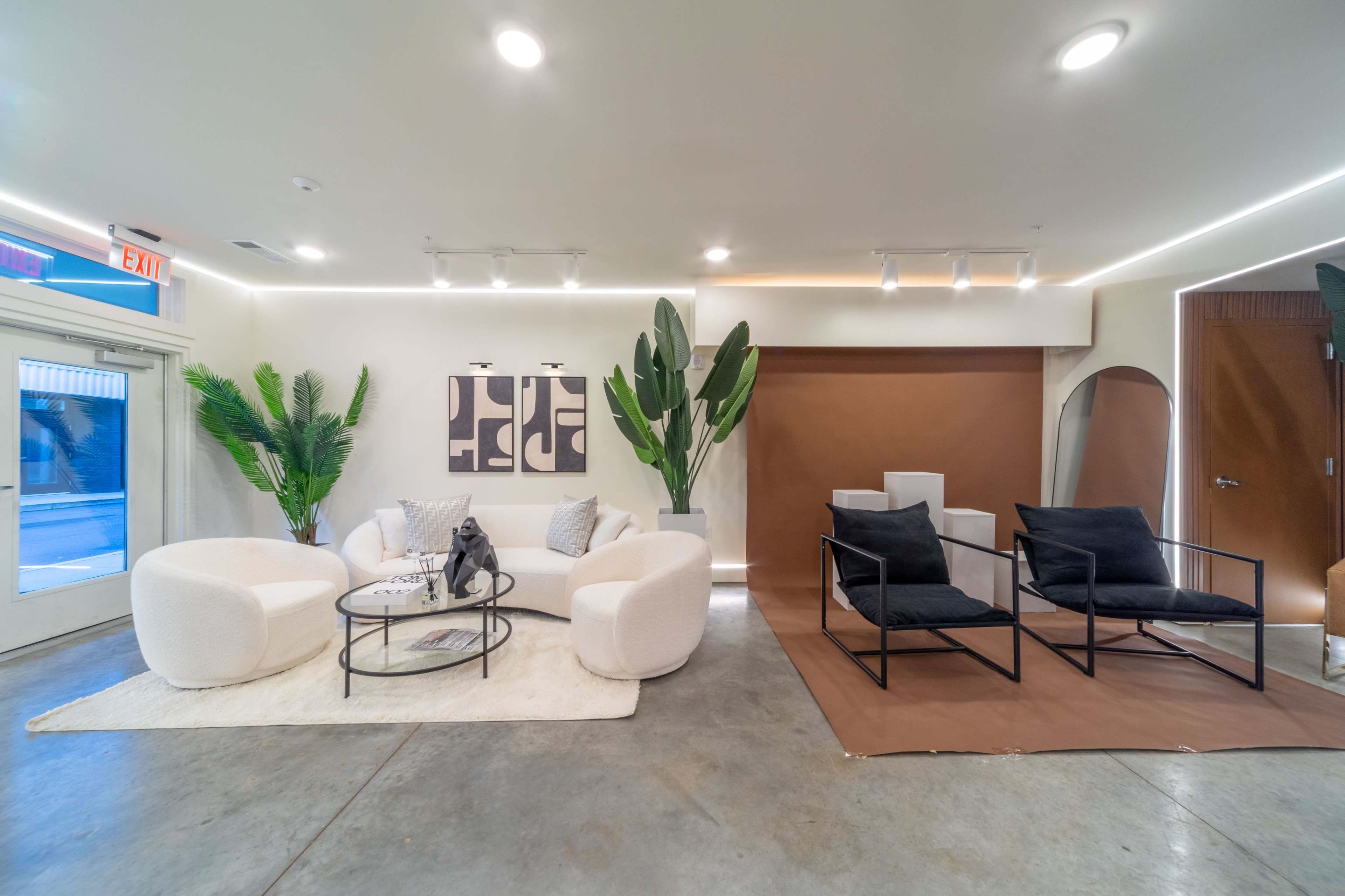 The image shows a modern interior space featuring a circular white seating arrangement, two black lounge chairs, large green plants, and minimalist artwork on the walls.