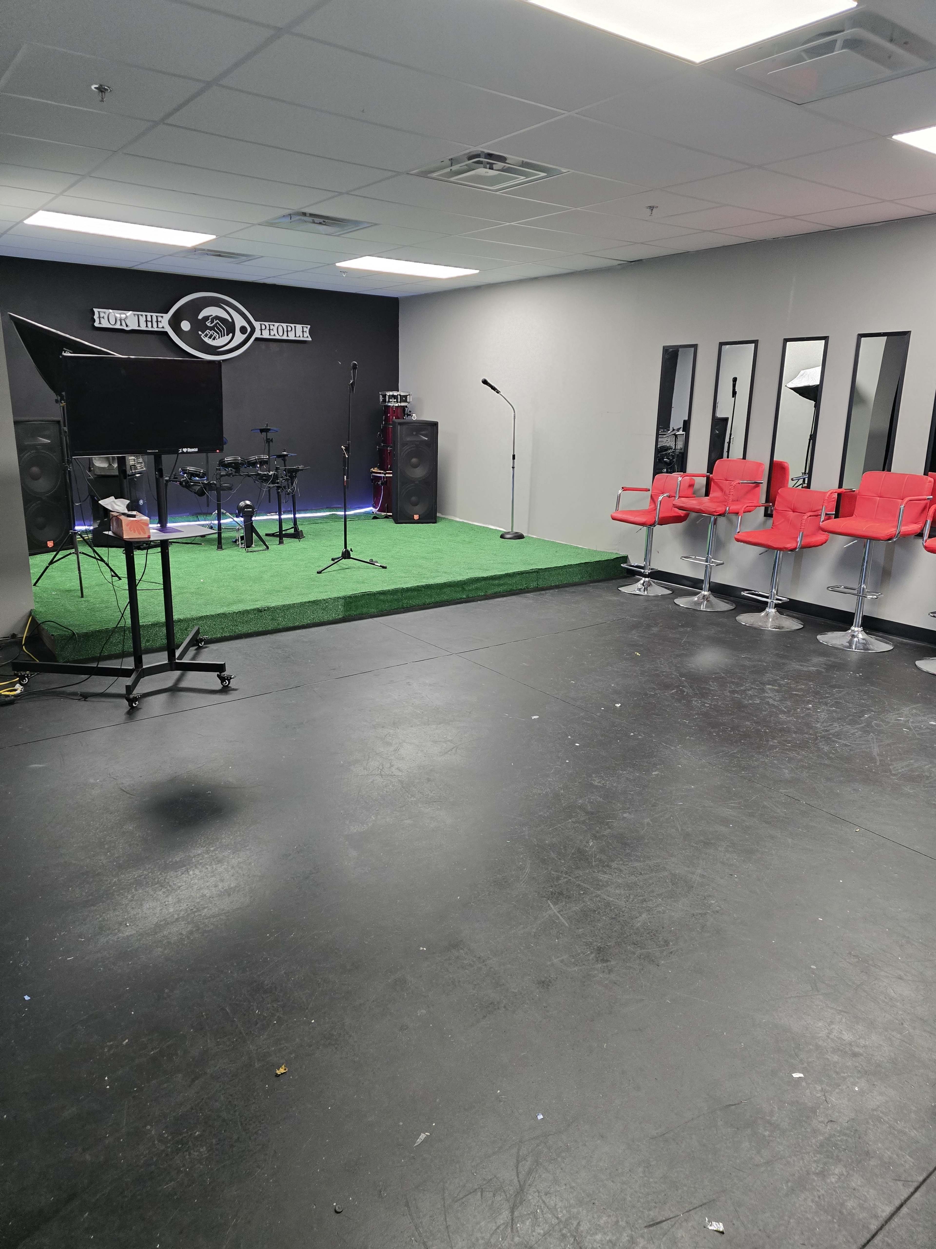 A music studio with a small stage, drum set, and red stools arranged near large mirrors.