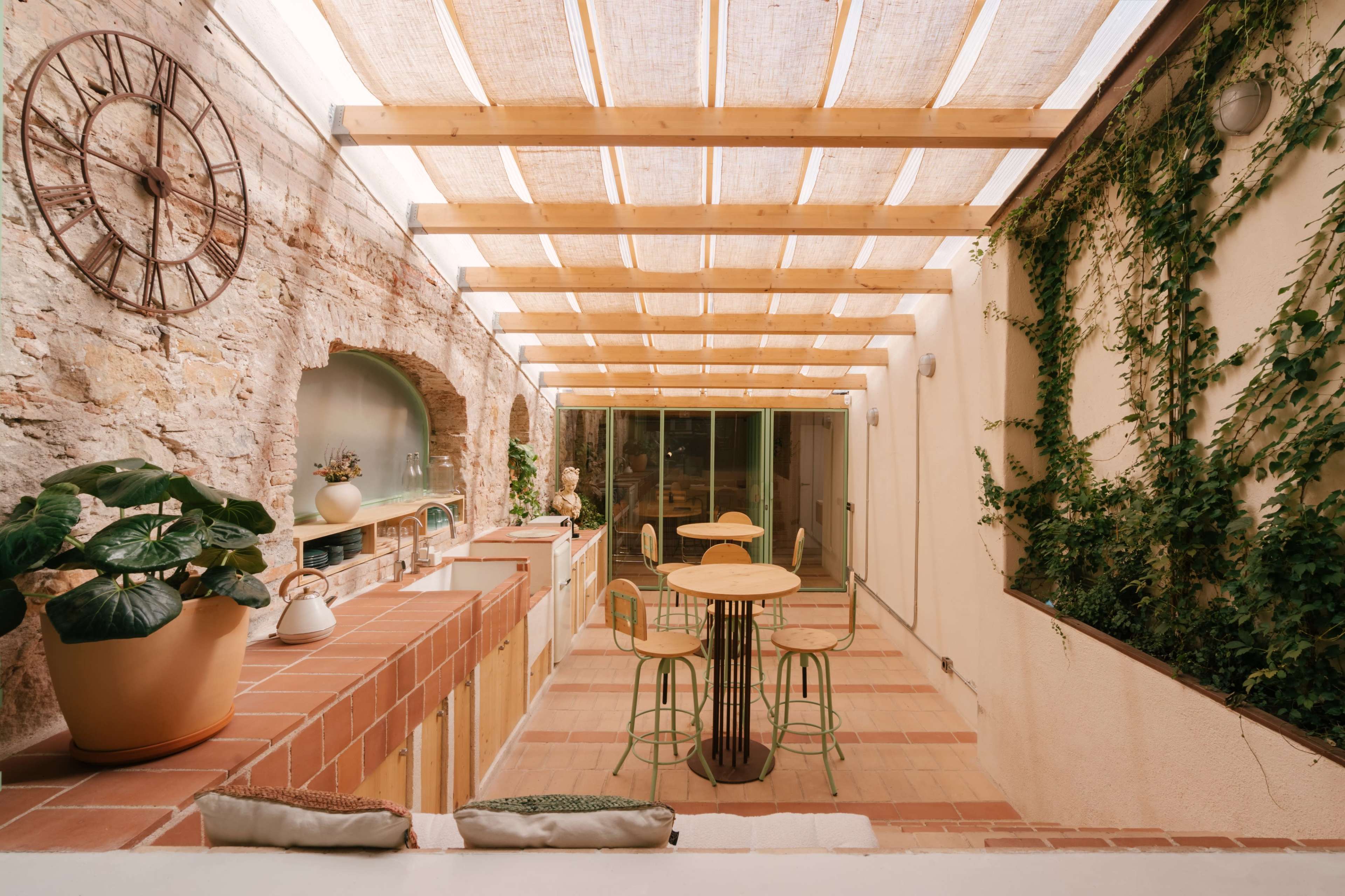 The image shows a modern outdoor dining area with a wooden trellis overhead, potted plants, a round table with four chairs, and a rustic stone wall.