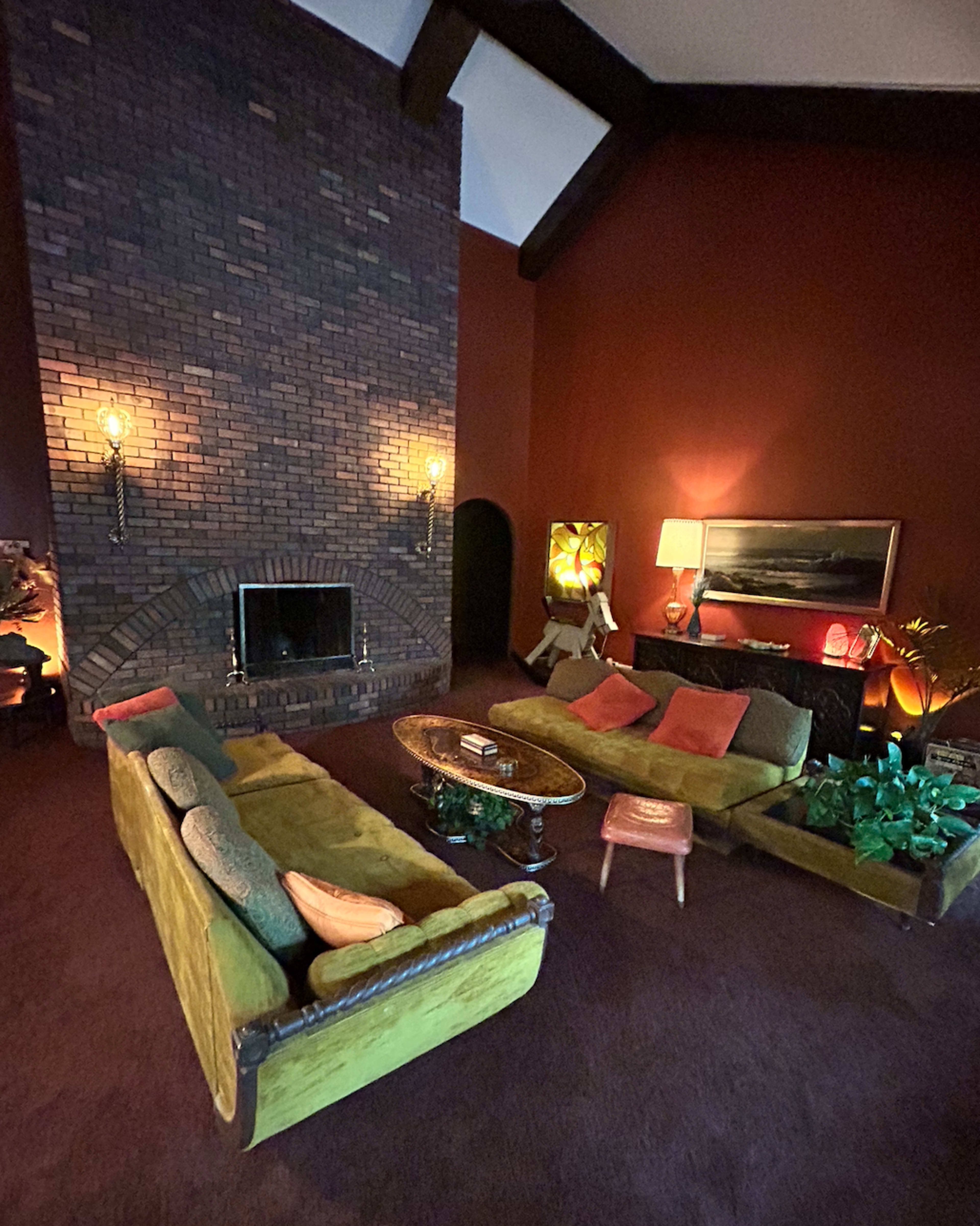 The image shows a retro living room with a brick fireplace, green sofas, and a circular coffee table surrounded by various decorative plants.