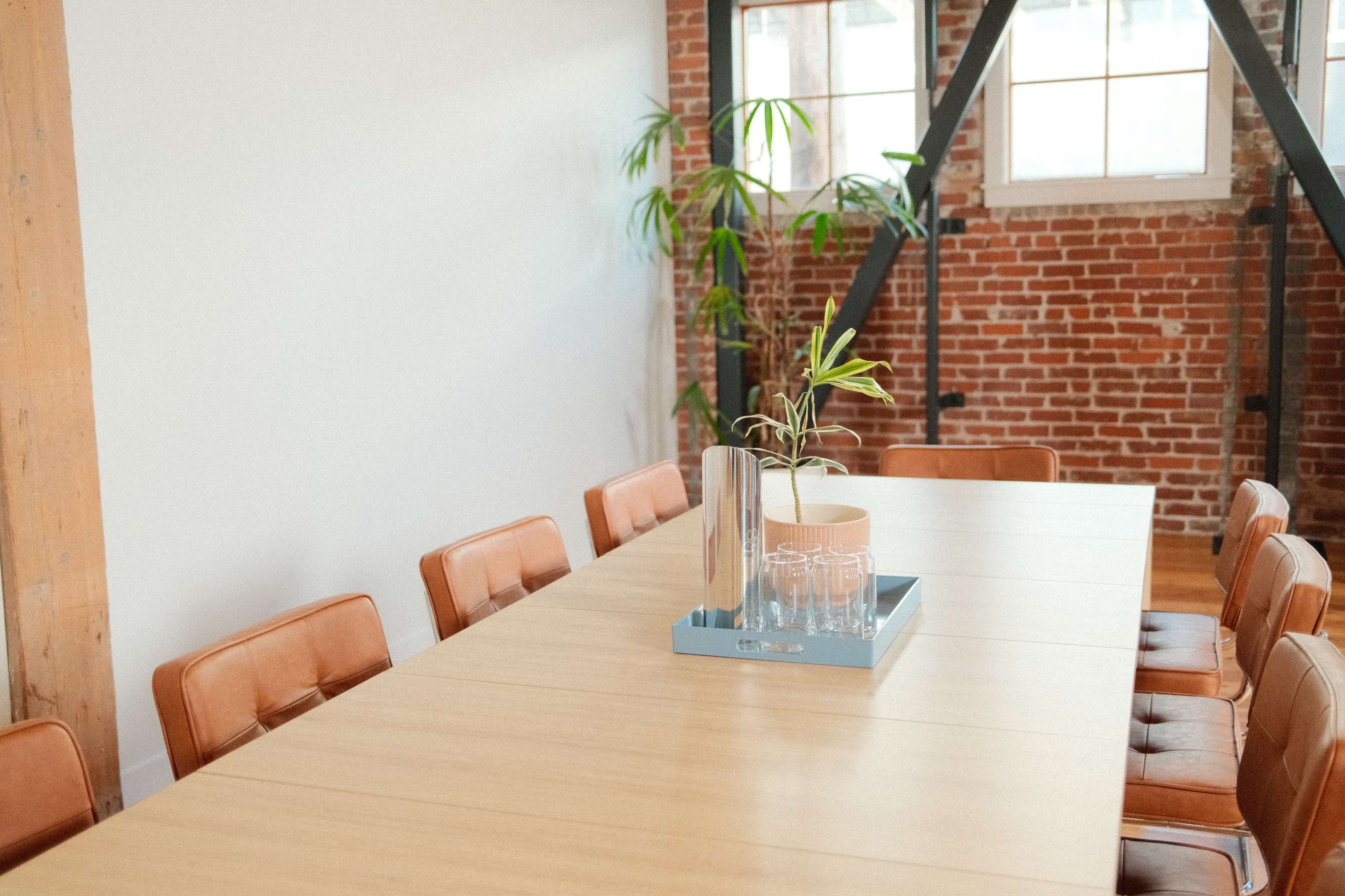 A long wooden conference table is surrounded by brown leather chairs in a room with exposed brick walls and natural light.