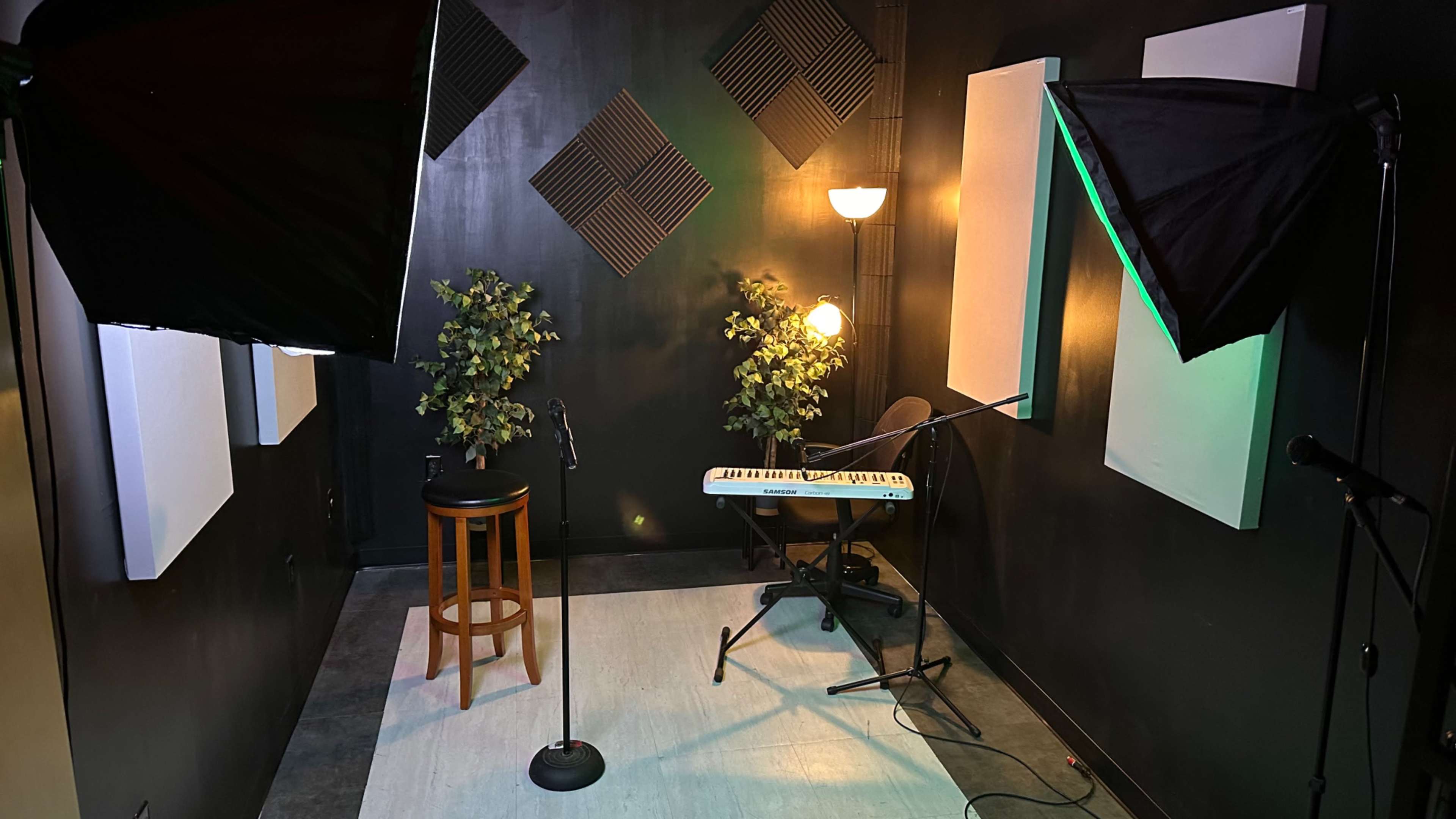 A small recording studio features microphone stands, a keyboard, a stool, and soft lighting against dark walls.