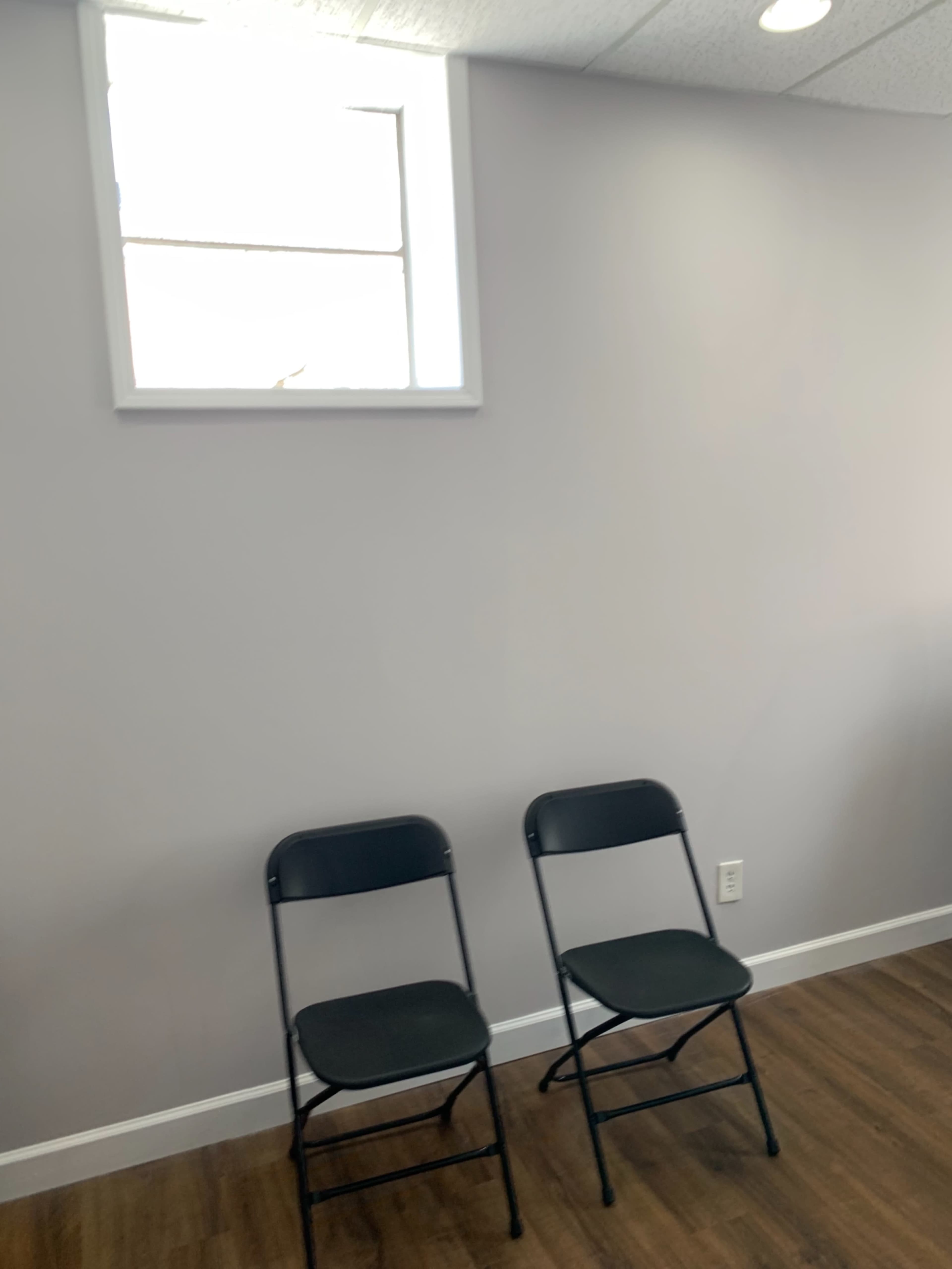 Two black folding chairs are positioned against a light gray wall beneath a small window.